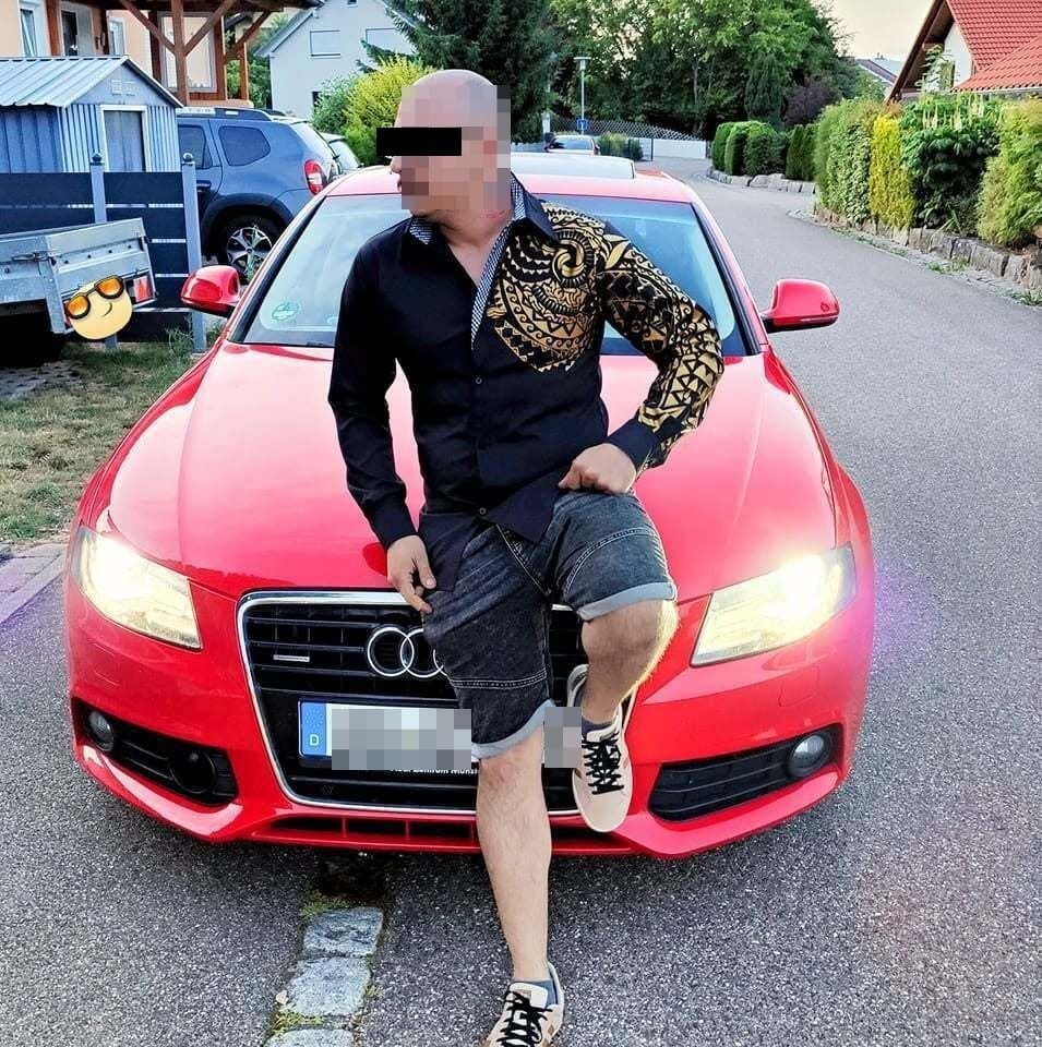 Ein Mann lehnt an einem roten Audi, sein Gesicht ist verpixelt und unkenntlich gemacht.