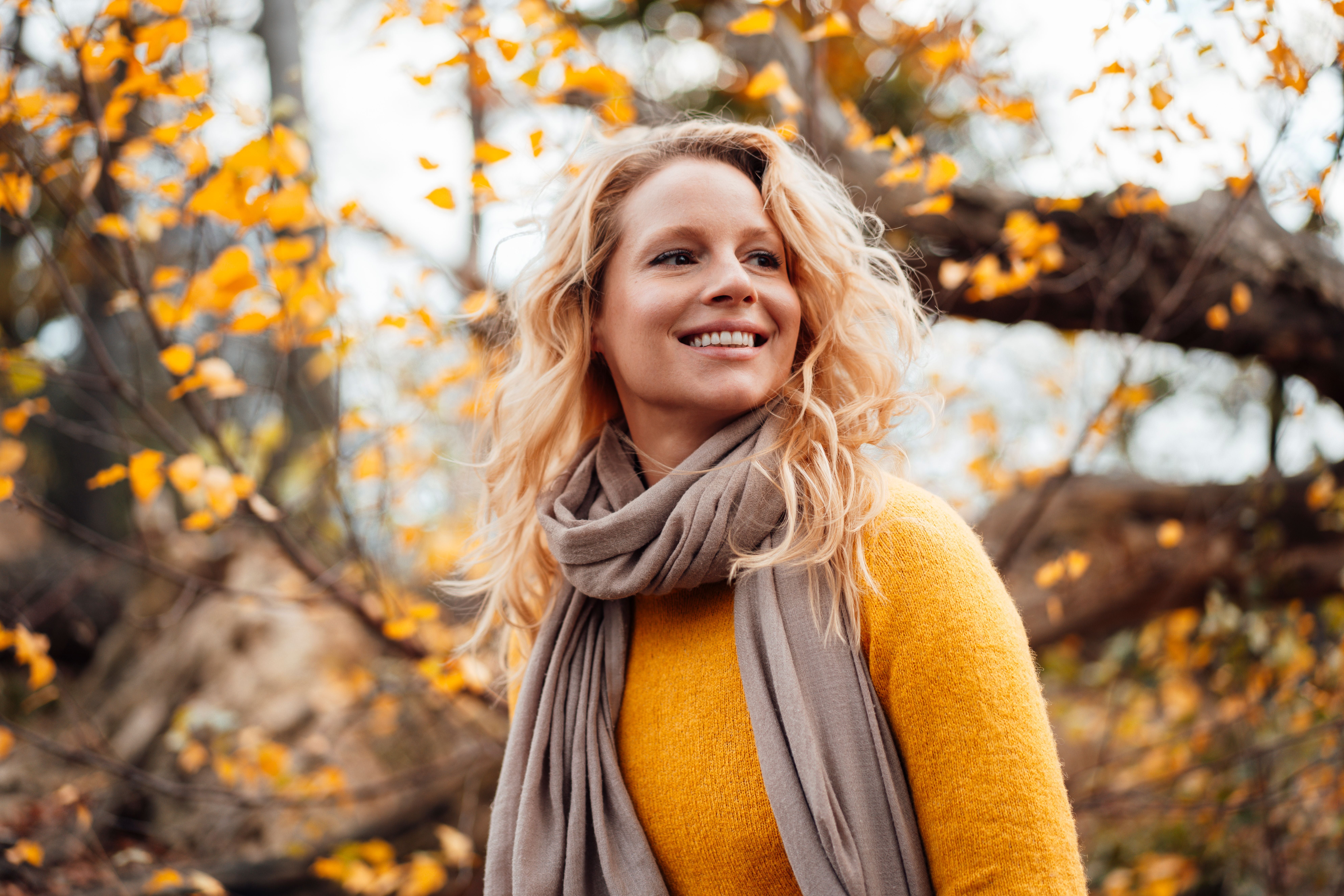 Frau mit blondem Haar und Schal lächelt im herbstlichen Park mit gelben Blättern.