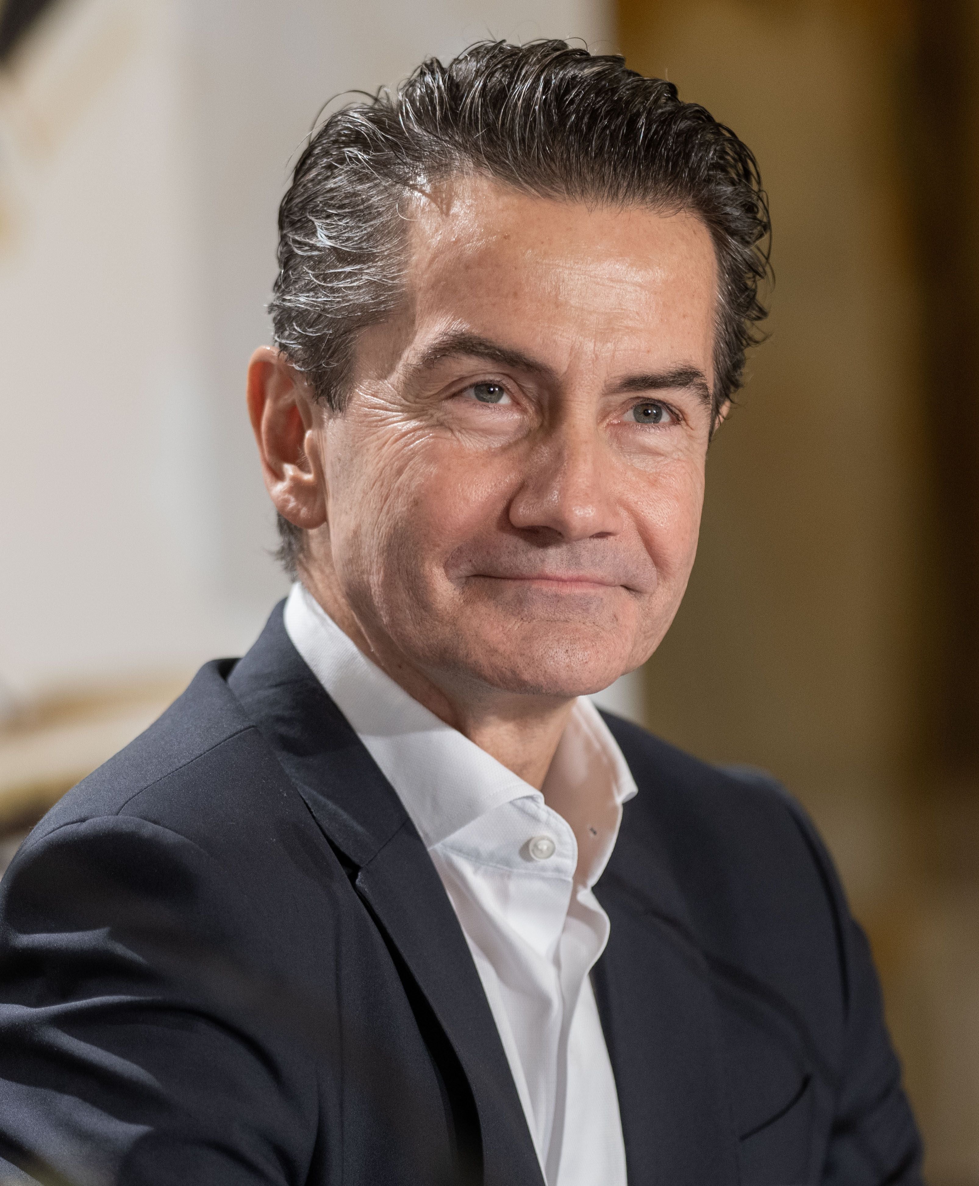 Ein Mann im Anzug sitzt und lächelt, Hintergrund ist unscharf.
