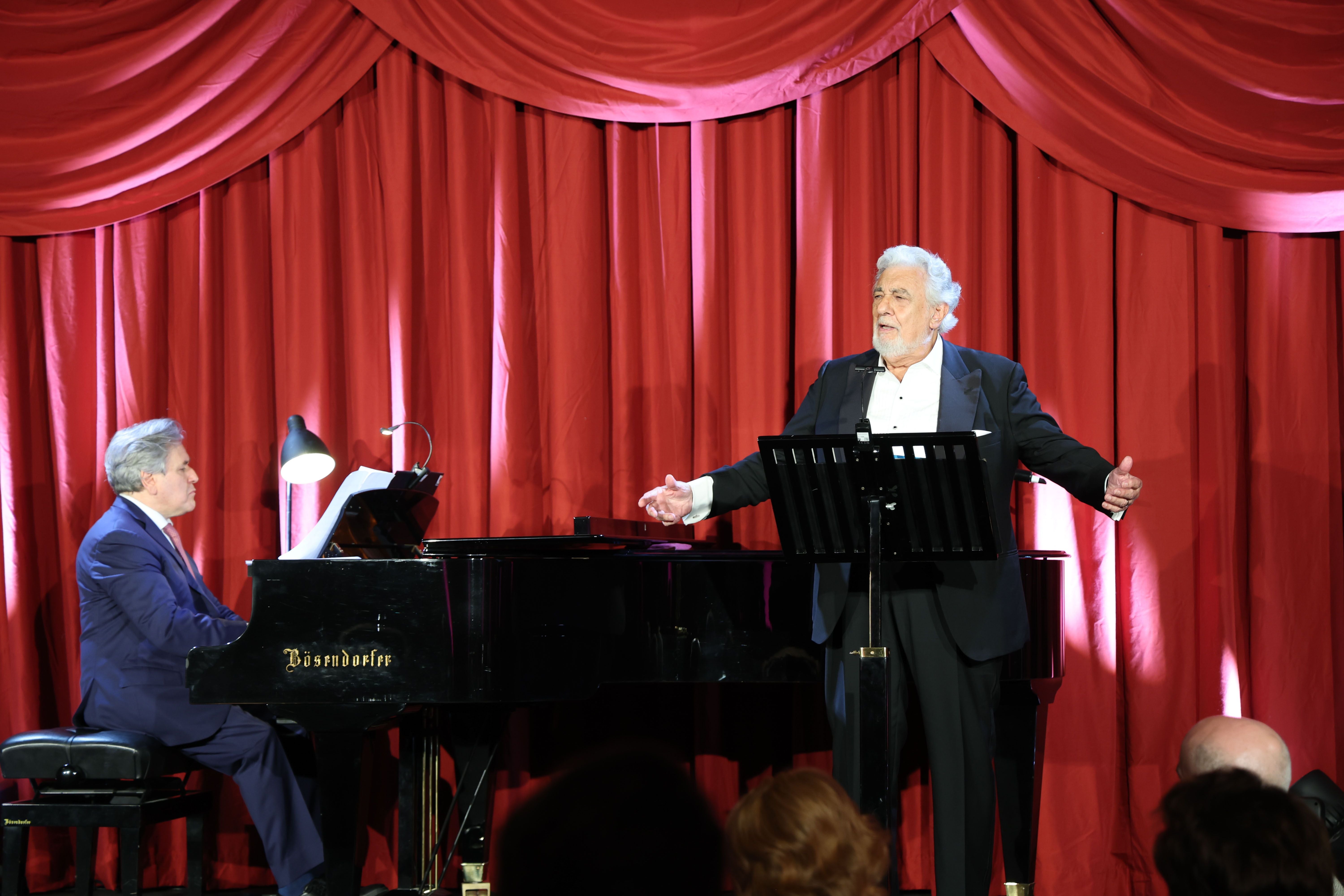 Ein Sänger singt neben einem Pianisten auf einer Bühne mit rotem Vorhang bei einer Gala.