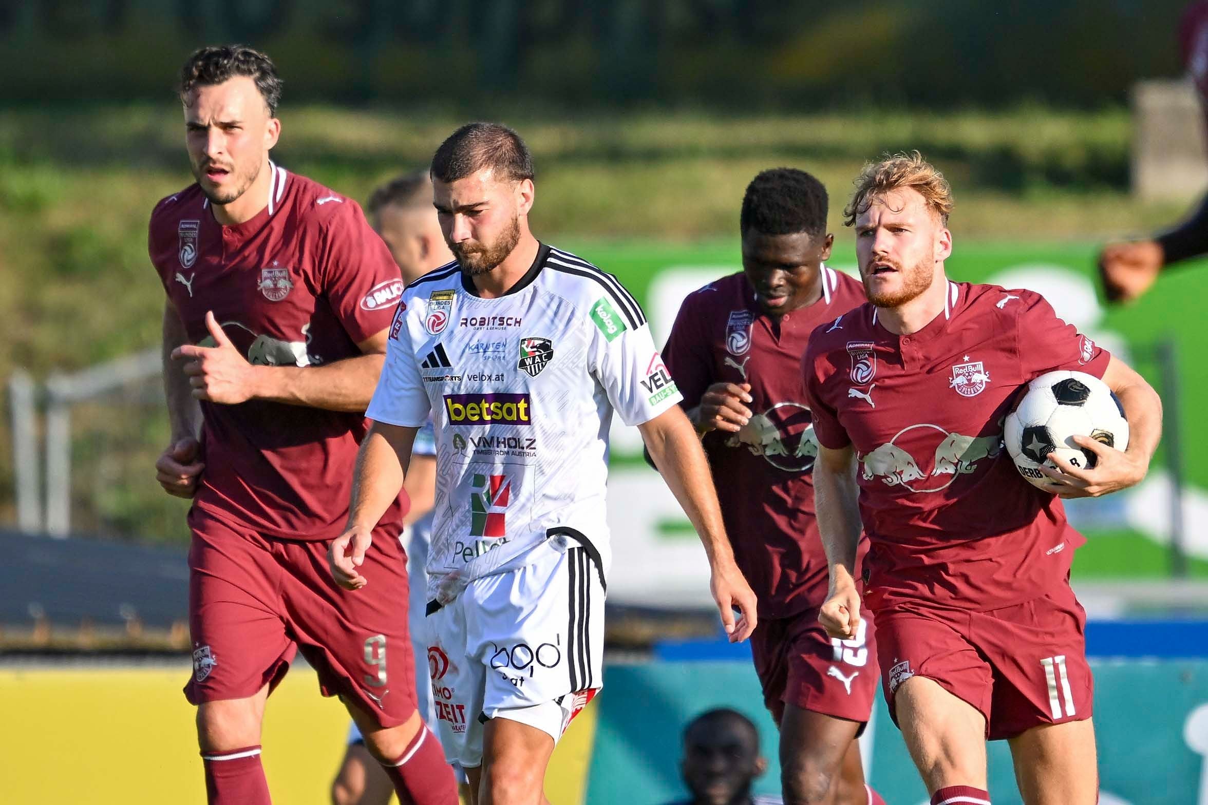 Vier Fußballspieler laufen auf dem Spielfeld, einer hält den Ball fest in der Hand.