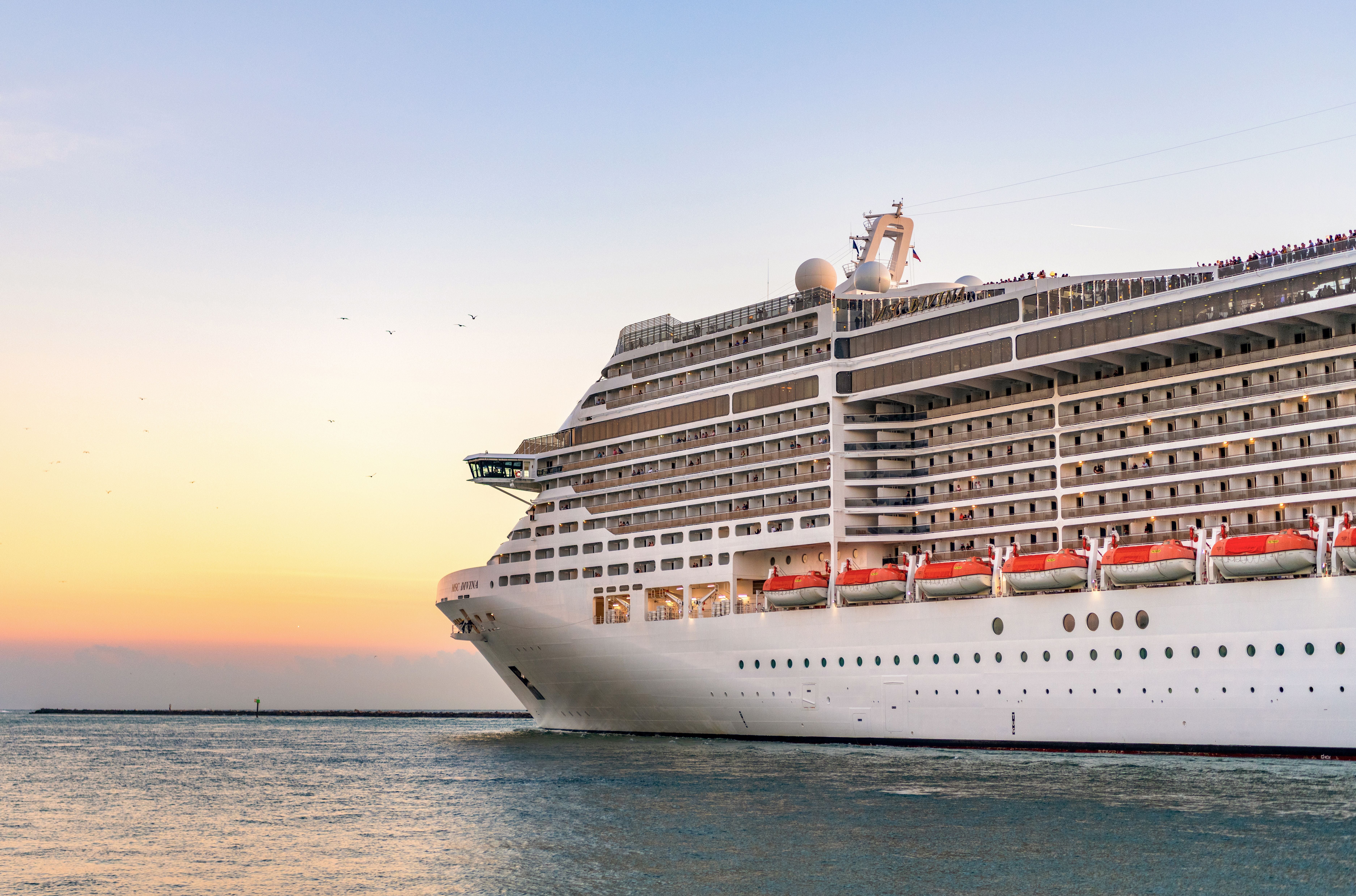 Großes Kreuzfahrtschiff verlässt den Hafen bei Sonnenuntergang, viele Balkonkabinen sichtbar.