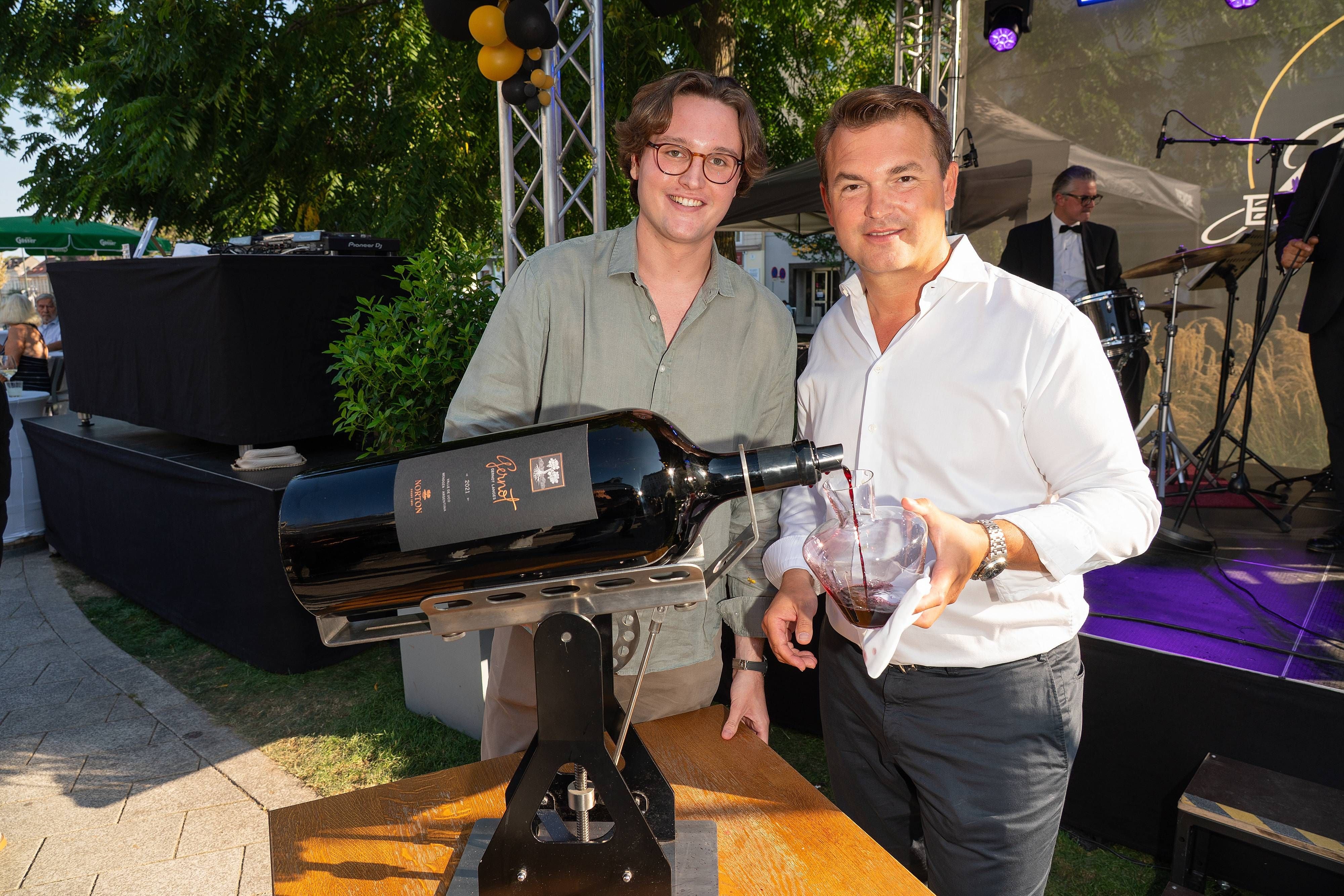 Zwei Männer schenken Wein aus einer riesigen Flasche in eine Karaffe bei einem Sommerfest ein.