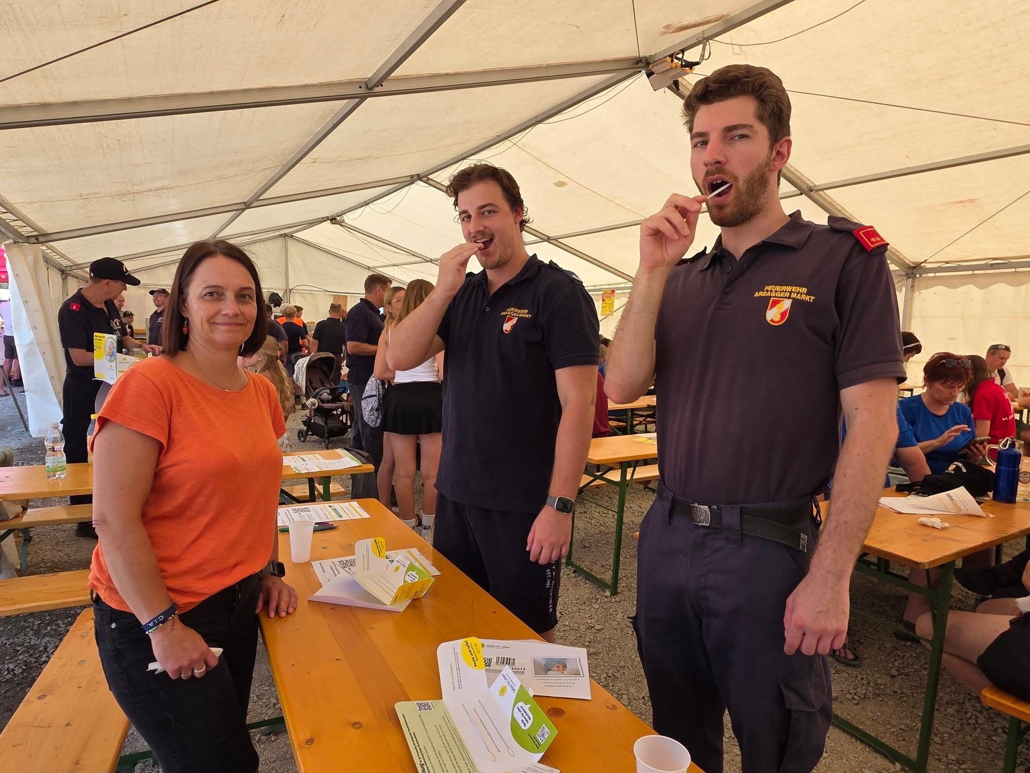 Drei Personen machen in einem Zelt einen Wangenabstrich, zwei tragen Feuerwehruniform.