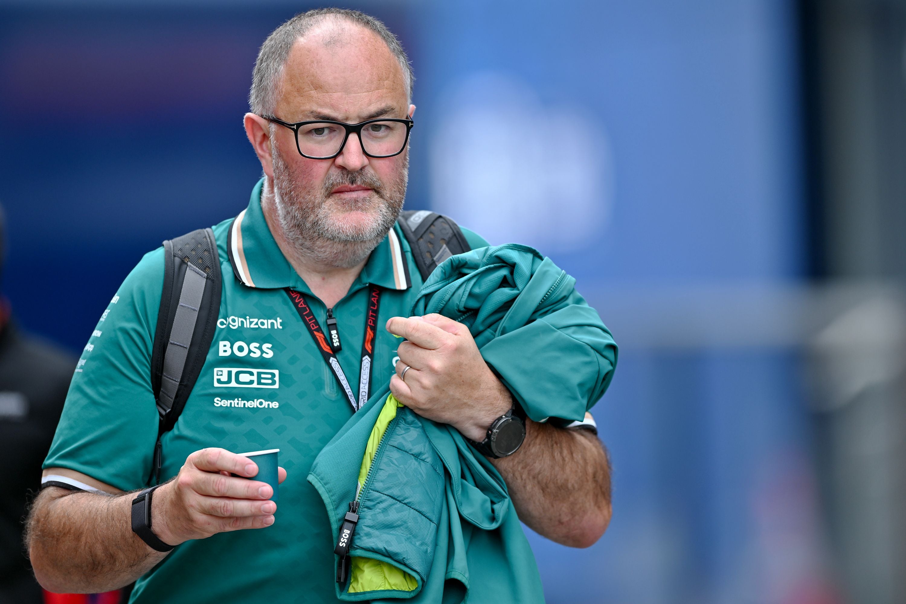 Ein Mann im Aston-Martin-F1-Team-Outfit hält Jacke und Becher, trägt Brille und Rucksack.