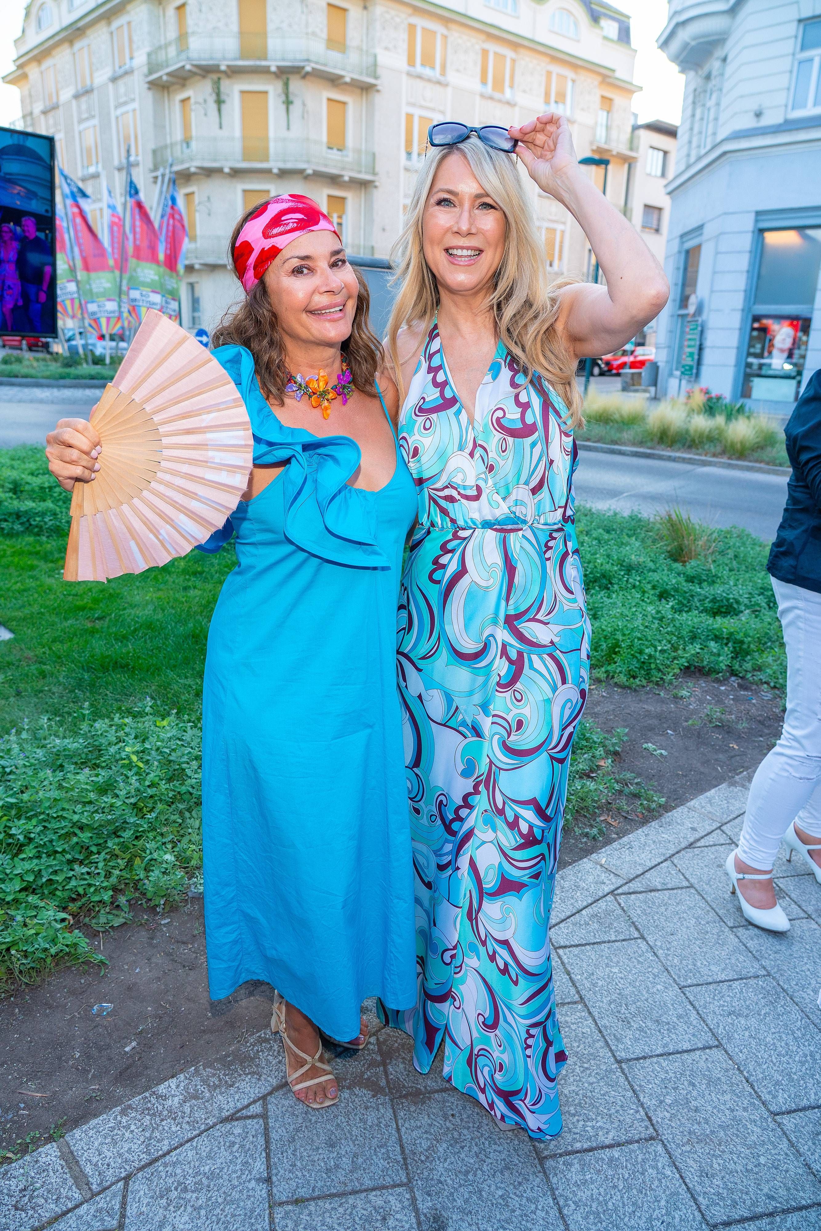 Zwei Frauen in bunten Sommerkleidern posieren lachend bei einer Outdoor-Veranstaltung.