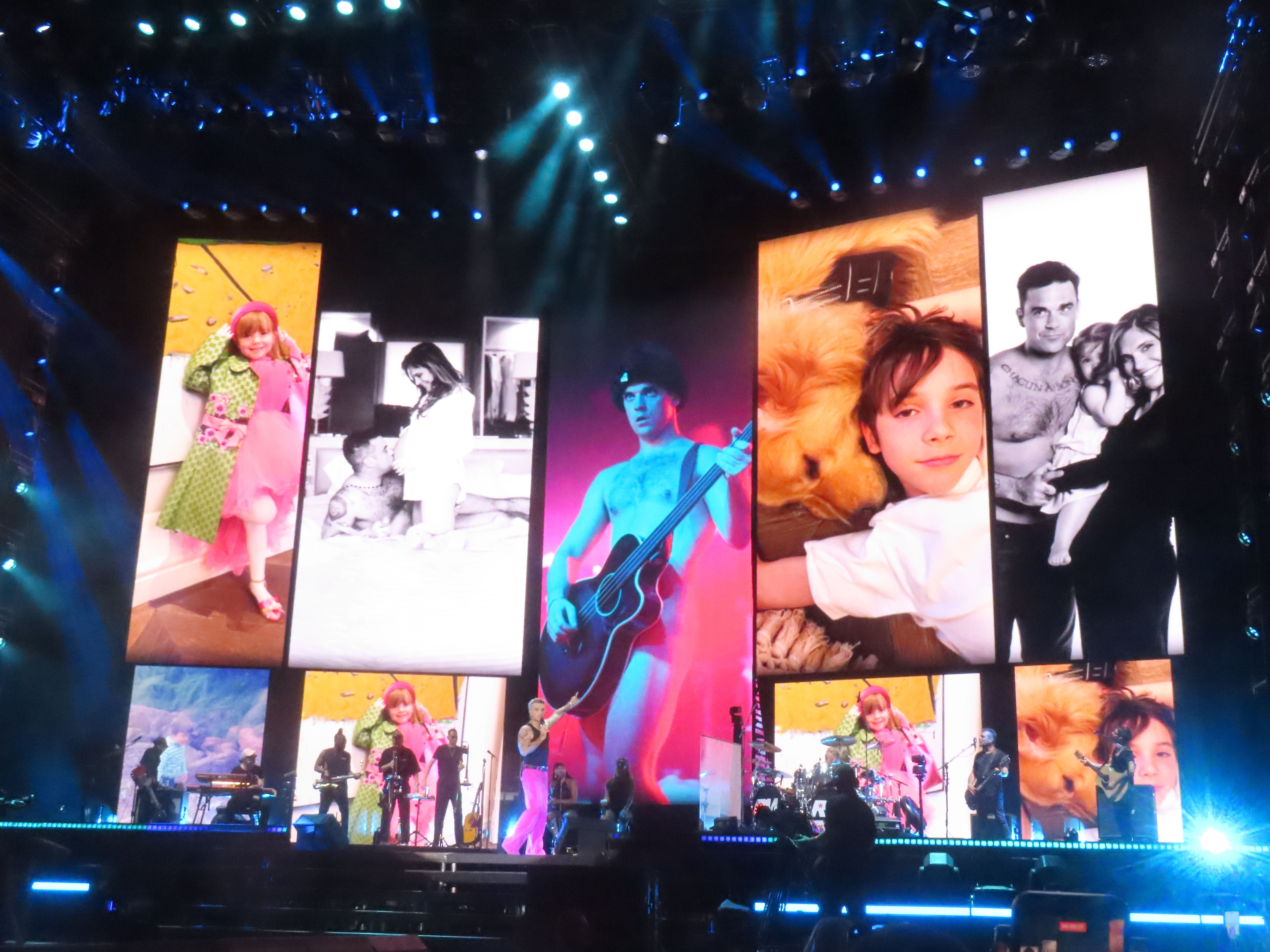 Robbie Williams und Band treten mit großen Familienfotos im Hintergrund auf Konzertbühne auf.