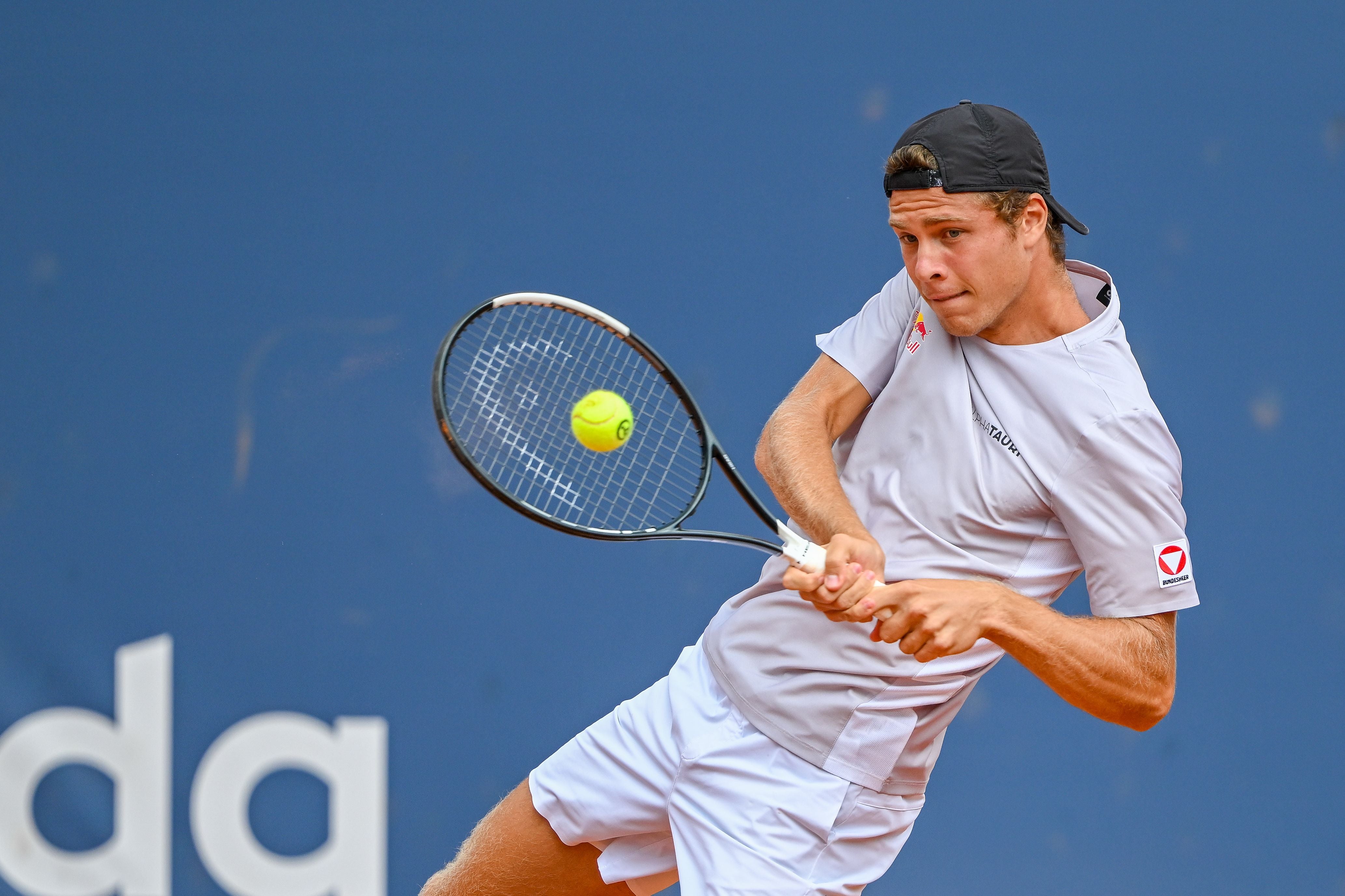 Ein Tennisspieler schlägt fokussiert einen Ball mit dem Schläger vor blauem Hintergrund.