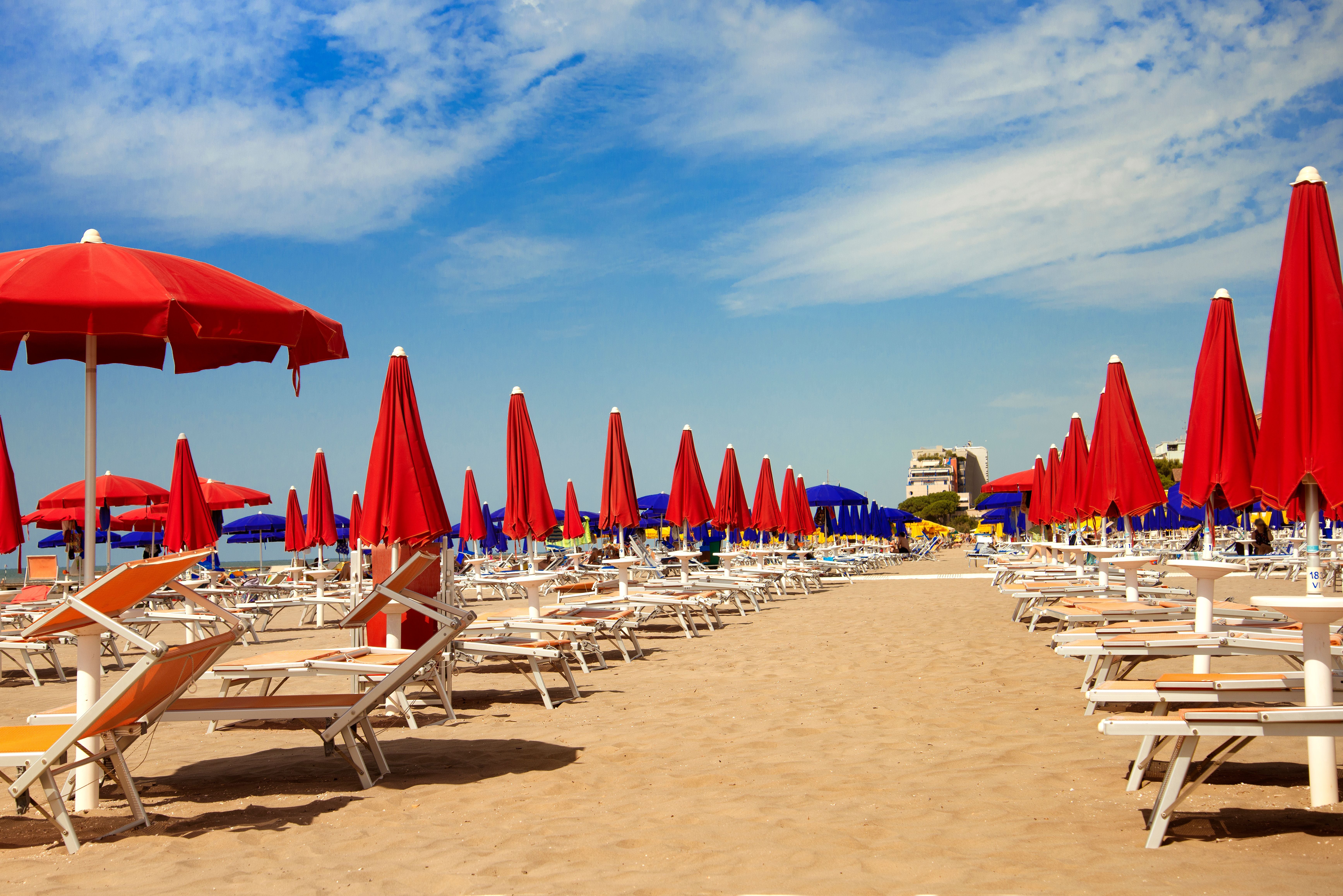 Reihen aus roten Sonnenschirmen und Liegen stehen am Sandstrand unter blauem Himmel.