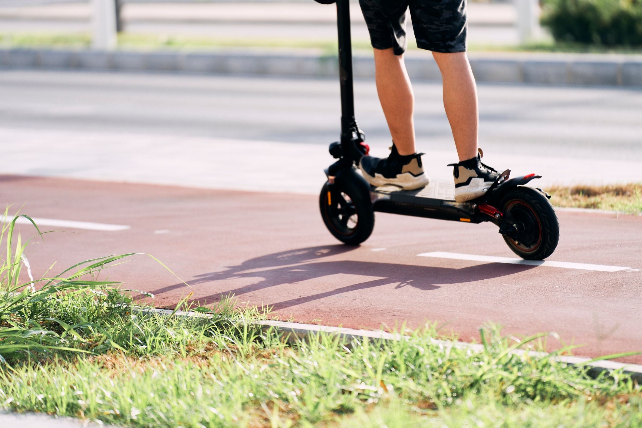 Person fährt auf einem Elektro-Tretroller auf einem Radweg, Grünfläche im Vordergrund.