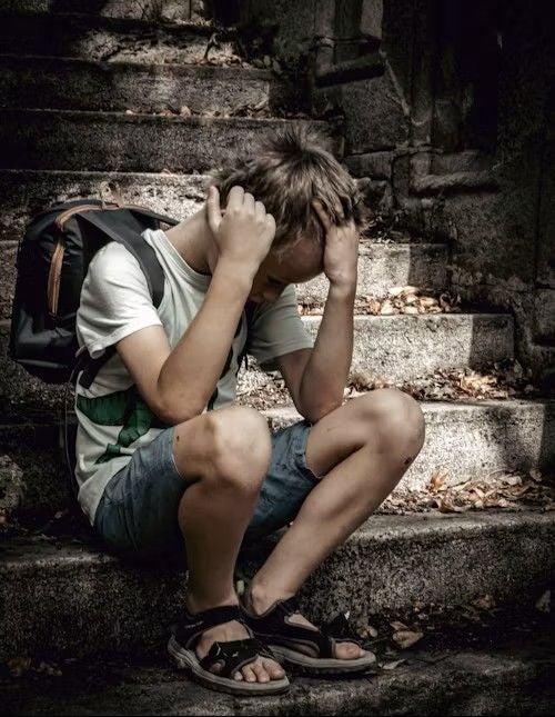Junge sitzt traurig auf einer Steintreppe, den Kopf in den Händen, mit einem Rucksack.