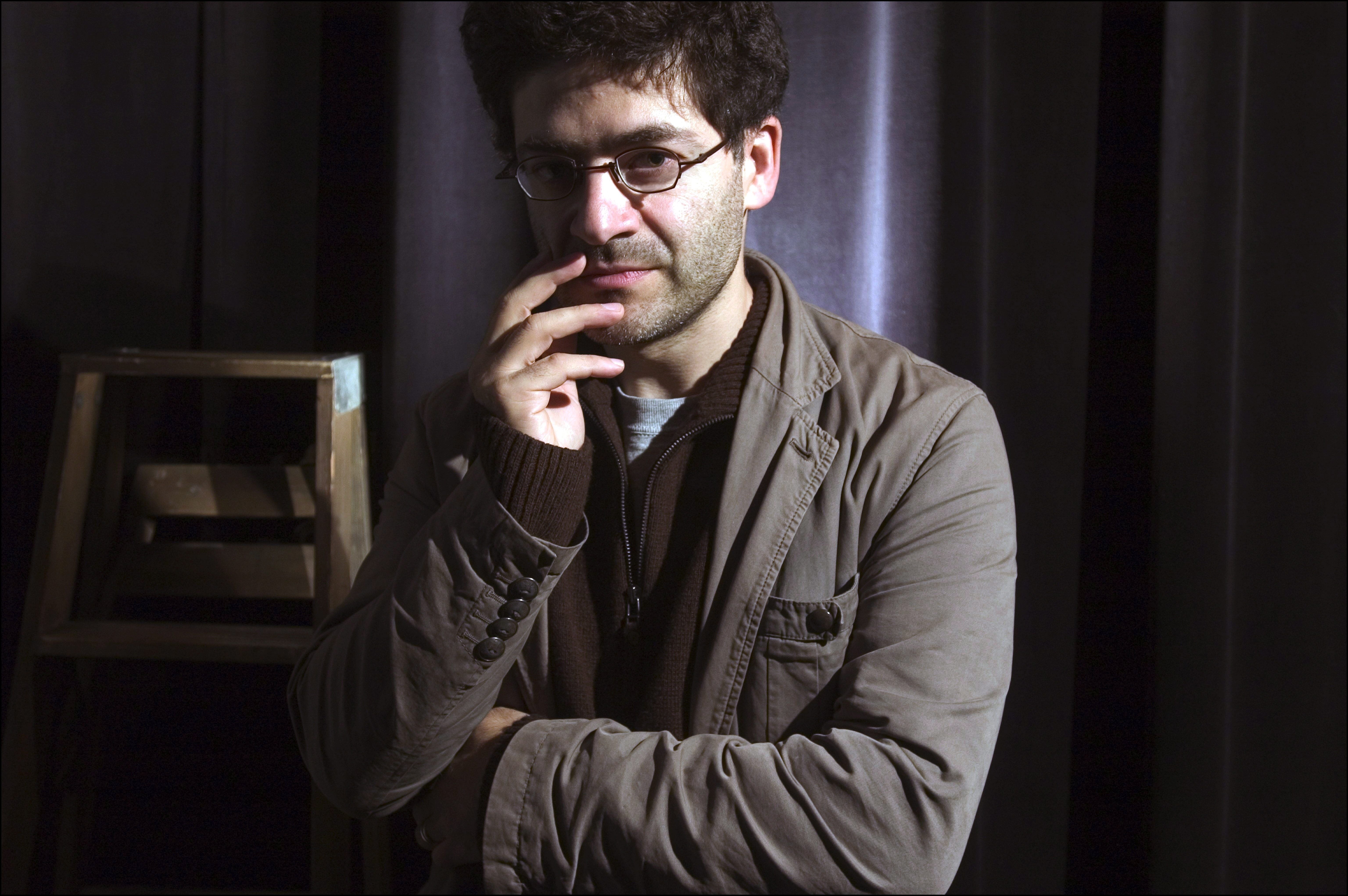 Ein Mann mit Brille steht nachdenklich vor einem dunklen Hintergrund und berührt sein Gesicht mit der Hand.