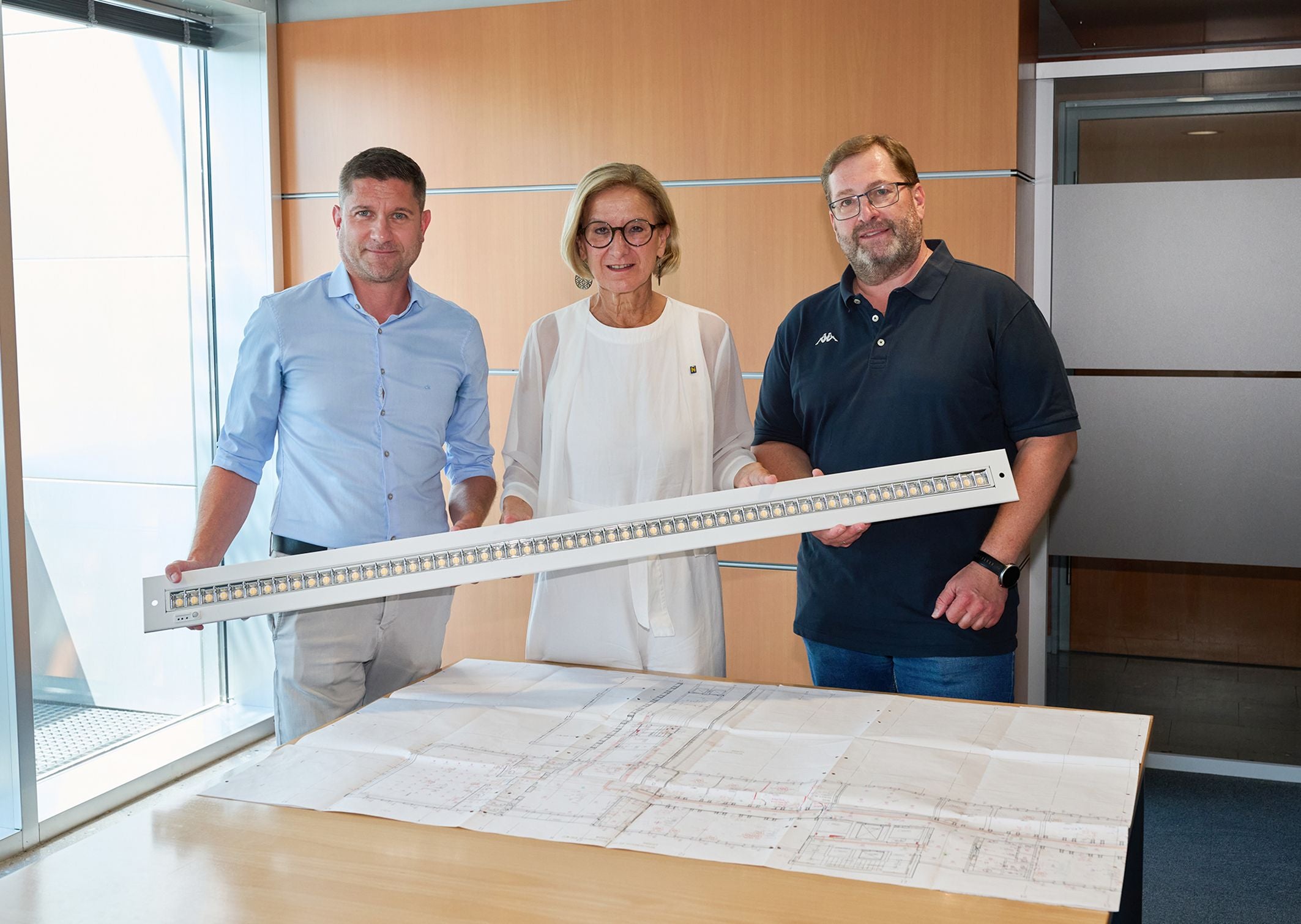 Drei Personen halten eine LED-Leuchte über einen Bauplan auf einem Tisch.