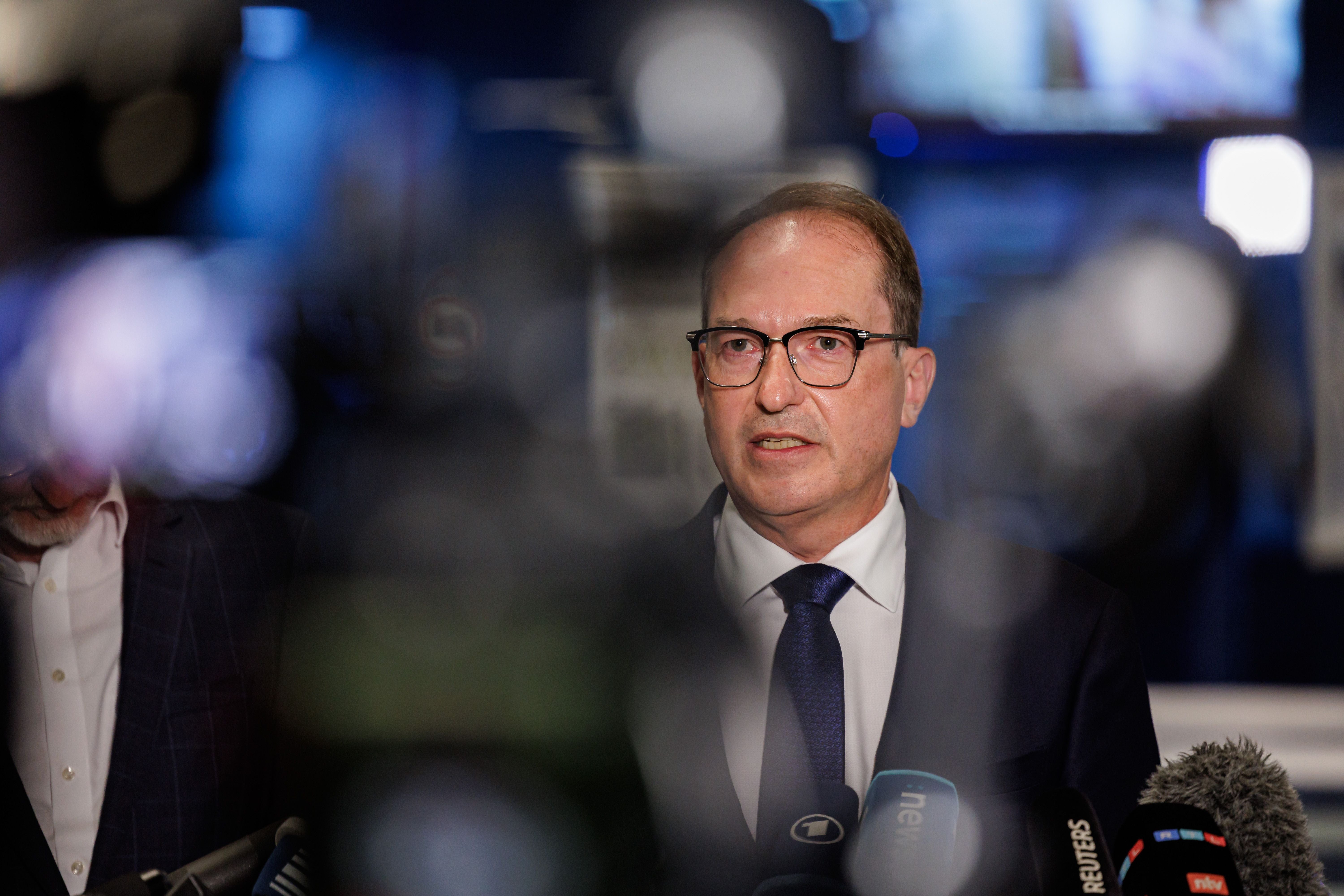 Ein Mann im Anzug spricht bei einer Pressekonferenz vor Mikrofonen und Kameras.