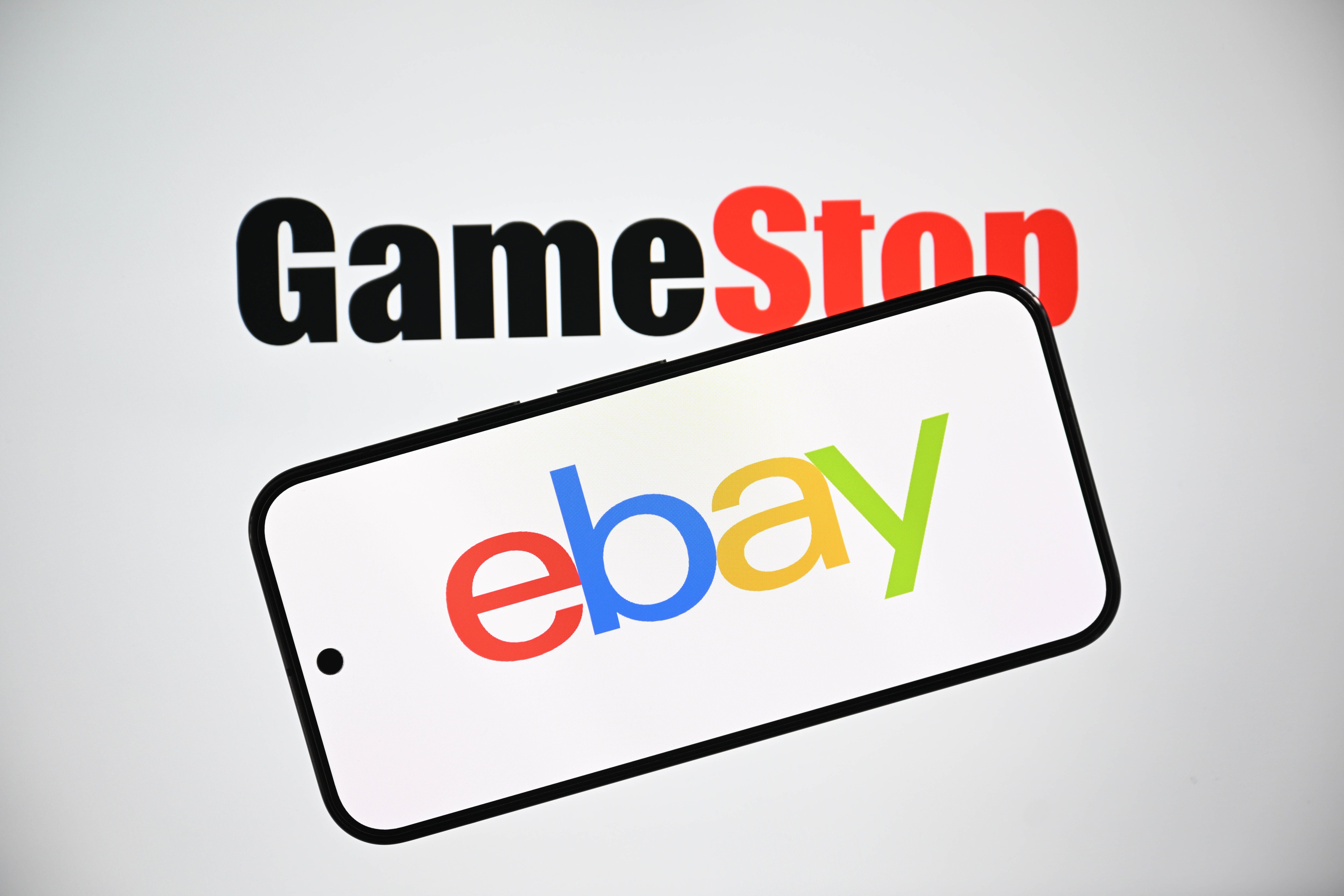 Das Gamestop-Logo im Hintergrund, das eBay-Logo auf einem Smartphone im Vordergrund.