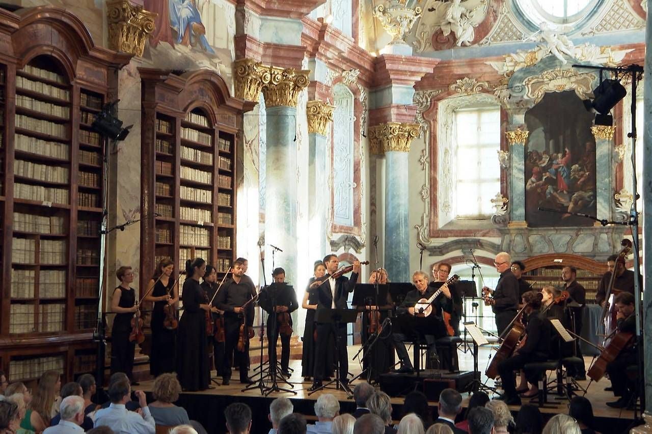 Konzert eines klassischen Orchesters in einem prunkvollen, historischen Saal mit Publikum.