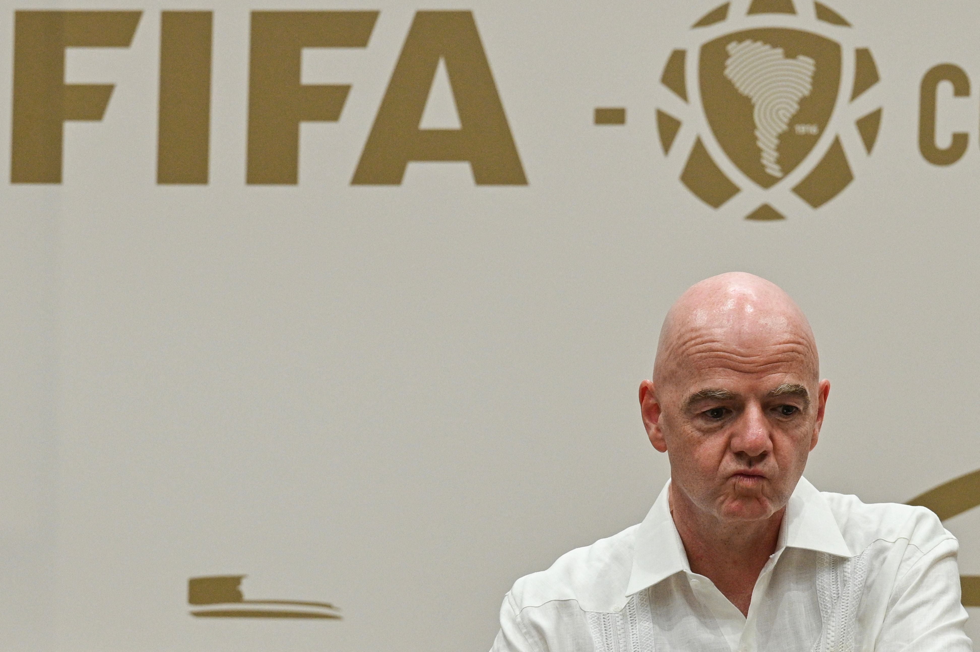 Ein Mann in weißem Hemd steht vor einer Wand mit großem FIFA-Logo und südamerikanischem Emblem.