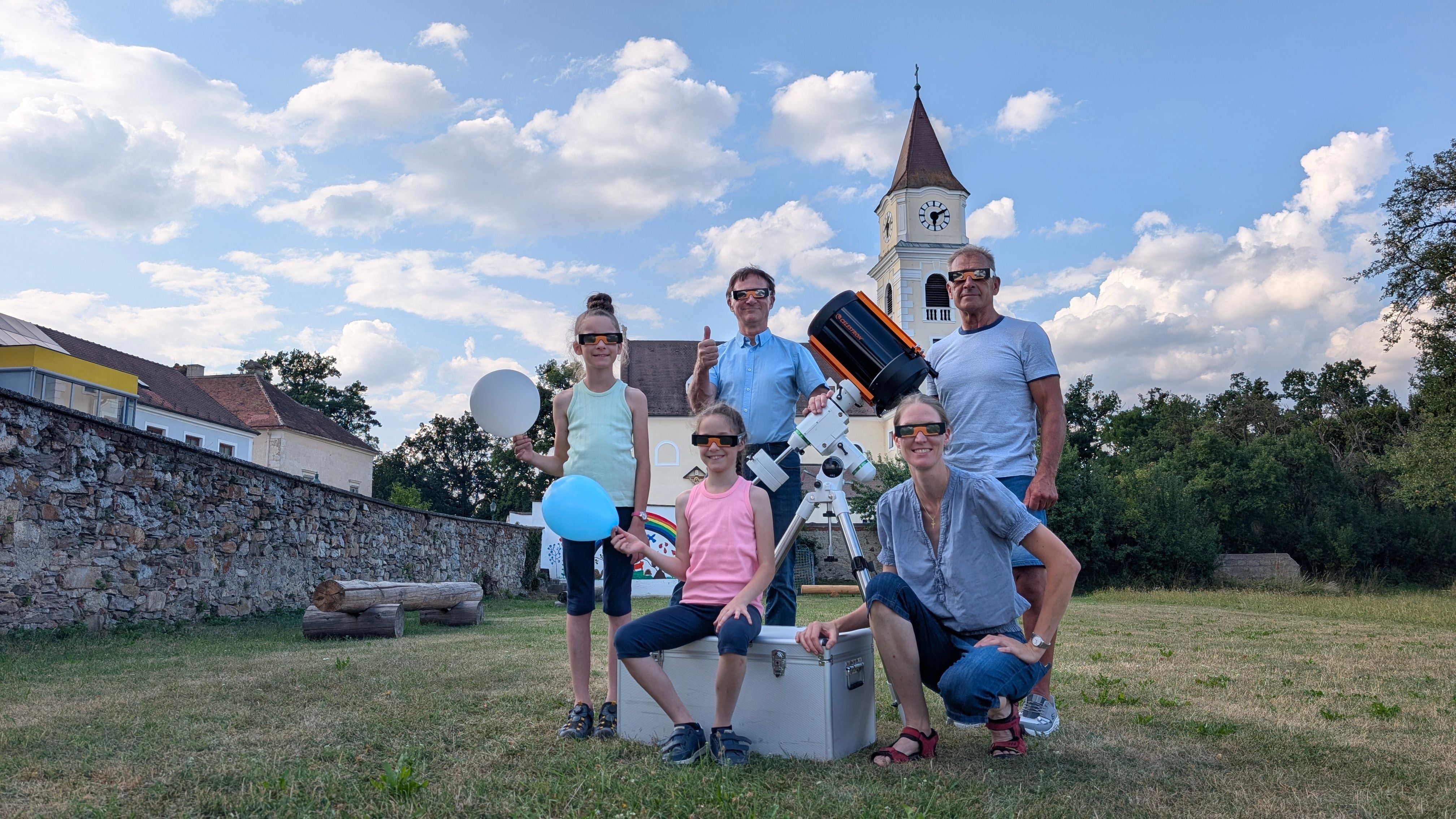 Fünf Personen mit Sonnenfinsternisbrillen und Teleskop im Freien vor einer Kirche.