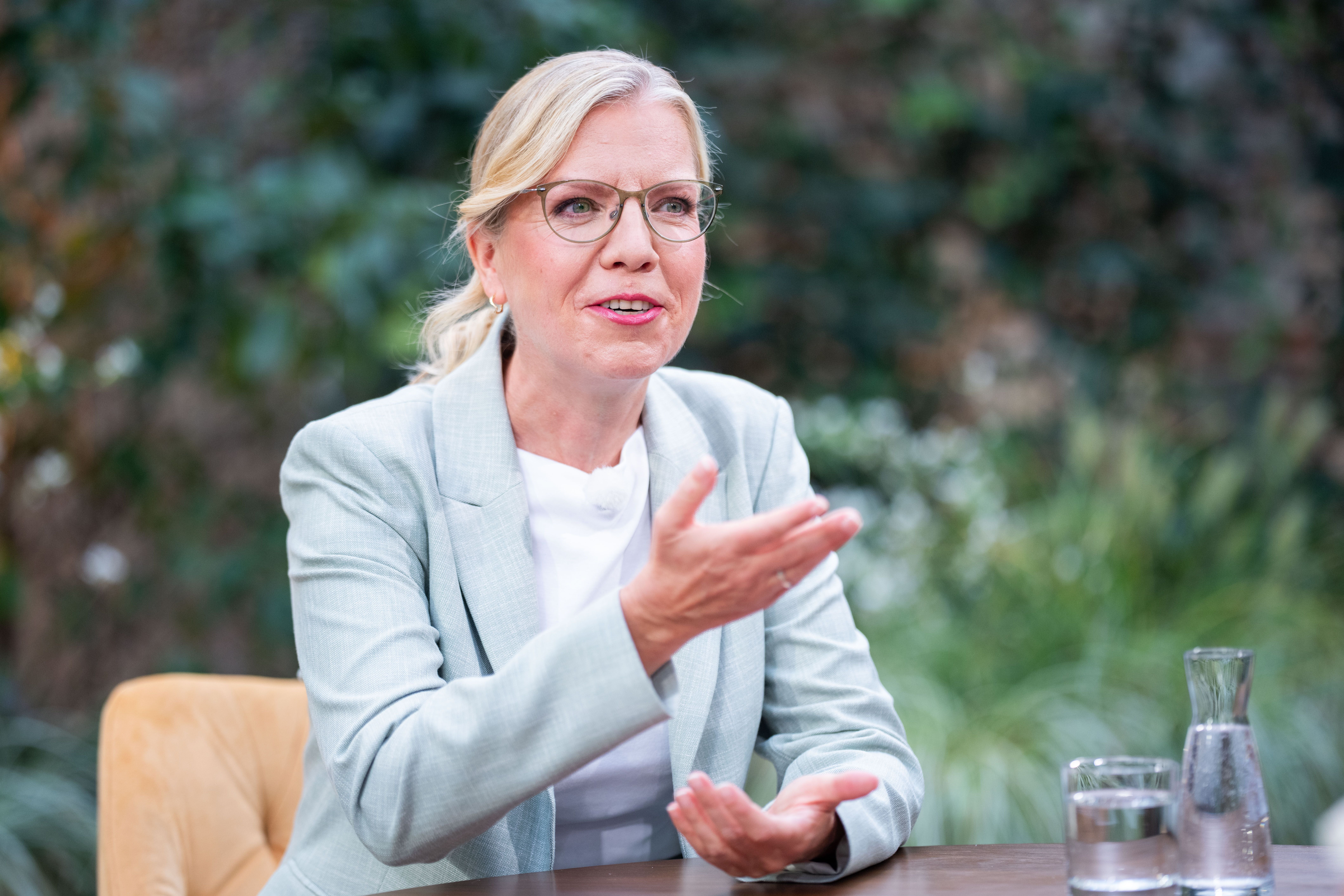 Frau mit Brille und hellem Blazer spricht gestikulierend an einem Tisch im Freien.