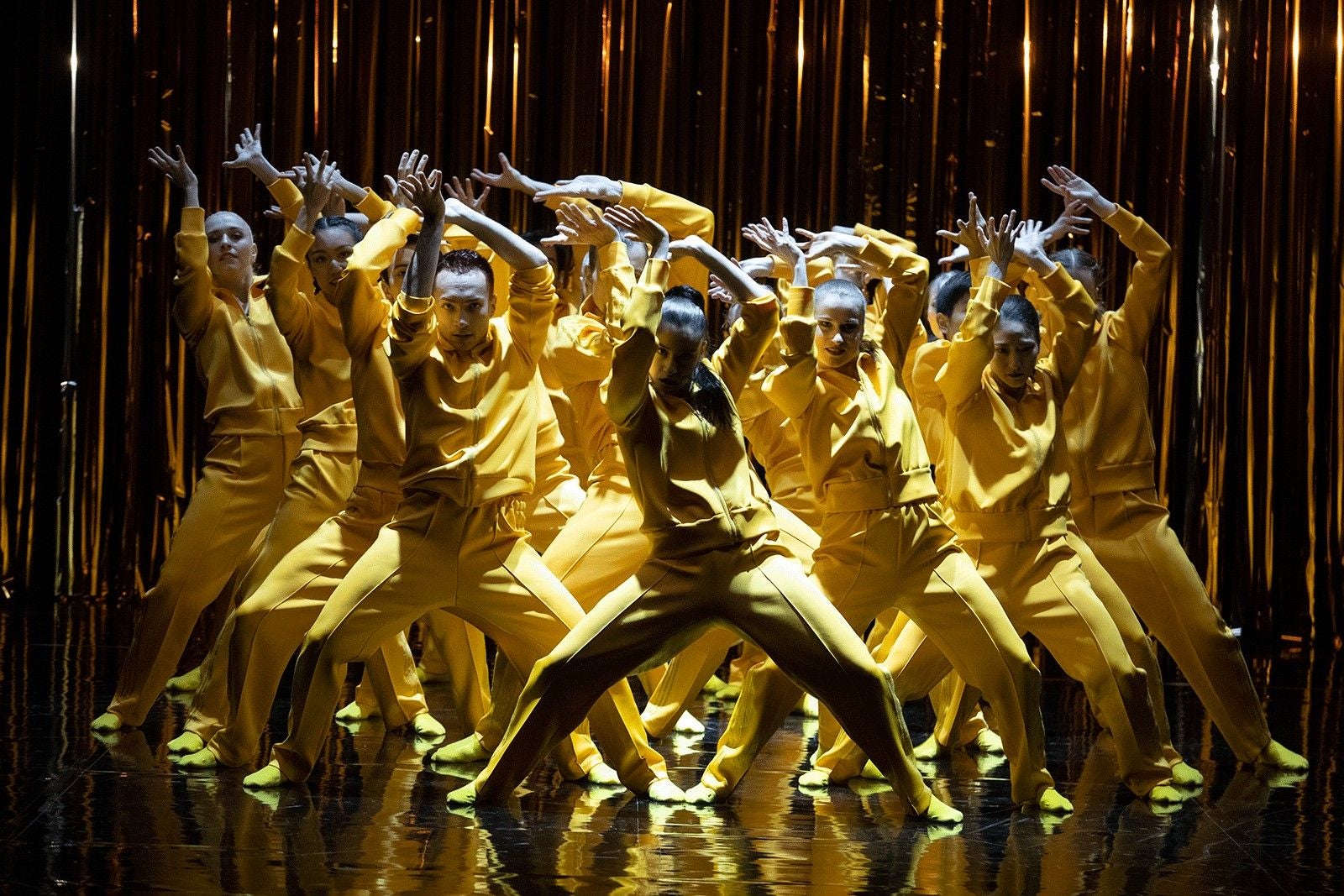 Tanzgruppe in gelben Outfits tanzt synchron auf Bühne mit goldenem Hintergrund.