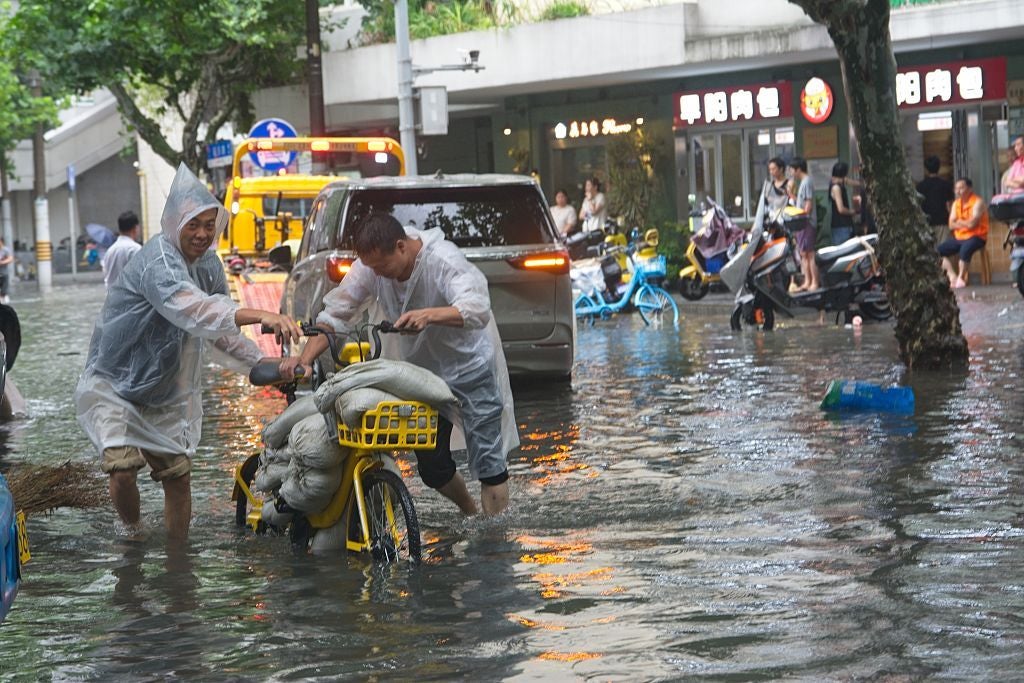 Zwei Männer in Regenmänteln schieben ein sandbagbeladenes Fahrrad durch überflutete Straßen.