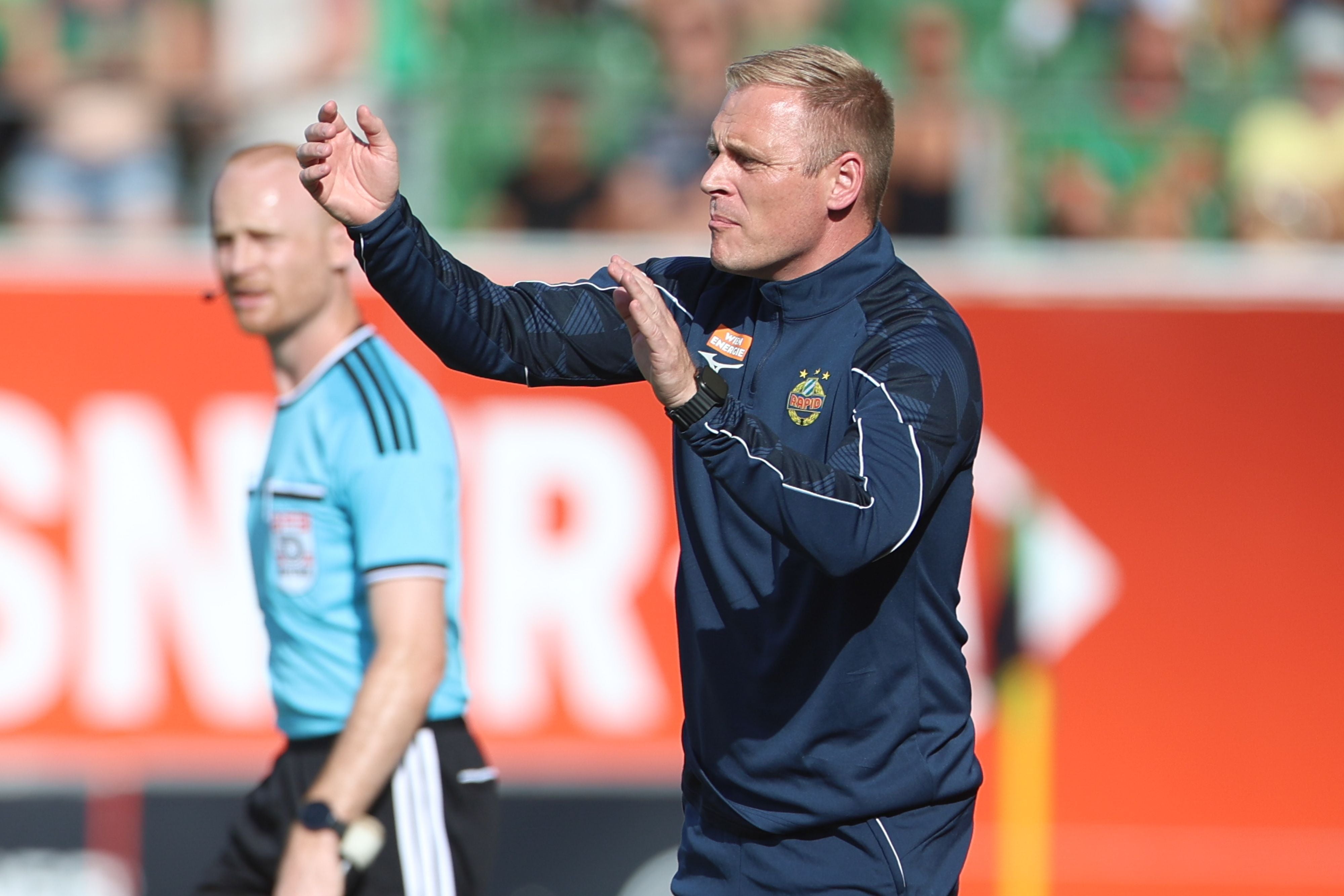 Trainer in Rapid-Trainingsanzug gestikuliert am Spielfeldrand, Schiedsrichter im Hintergrund.
