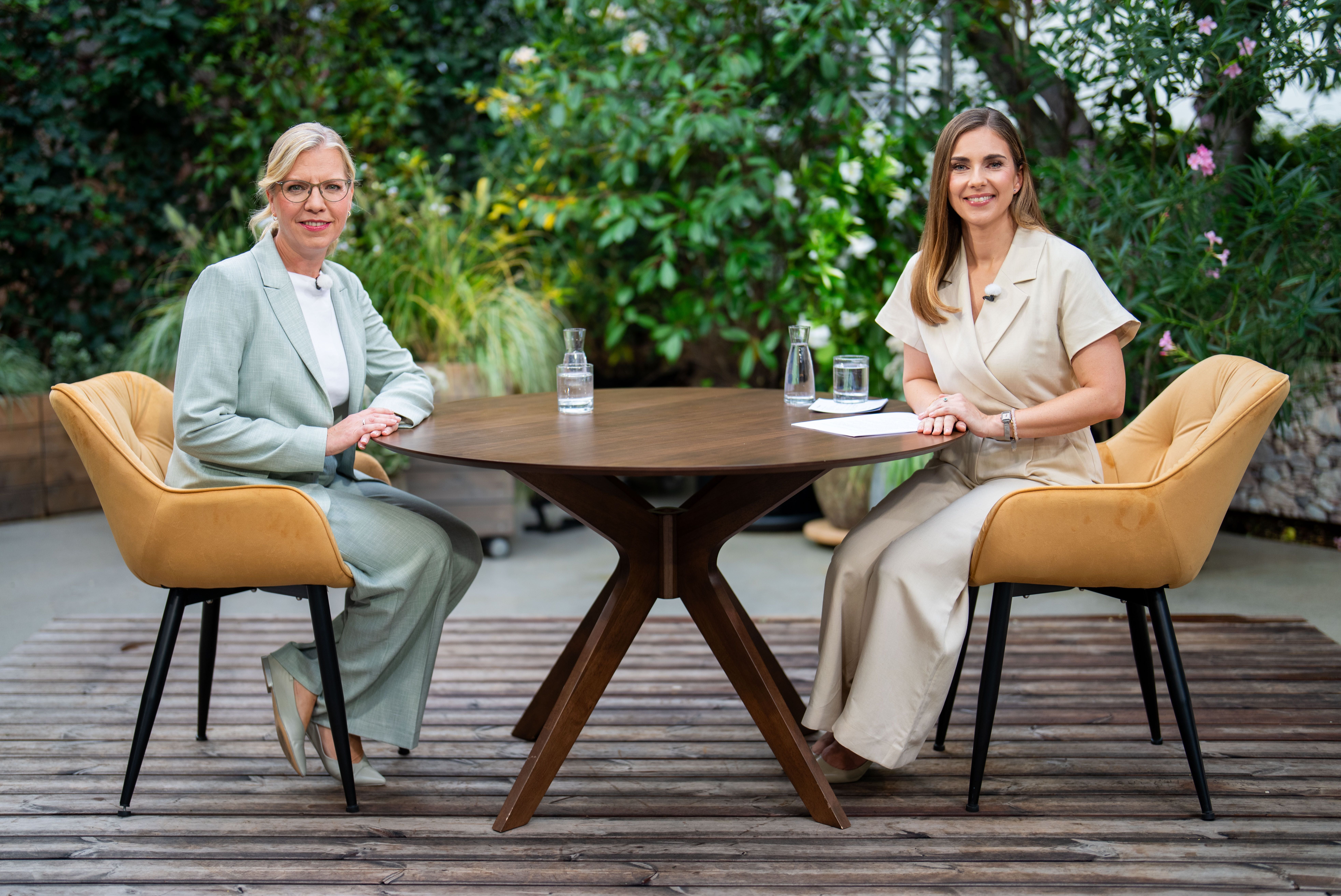 Zwei Frauen sitzen einander an einem runden Holztisch im Freien gegenüber und lächeln.