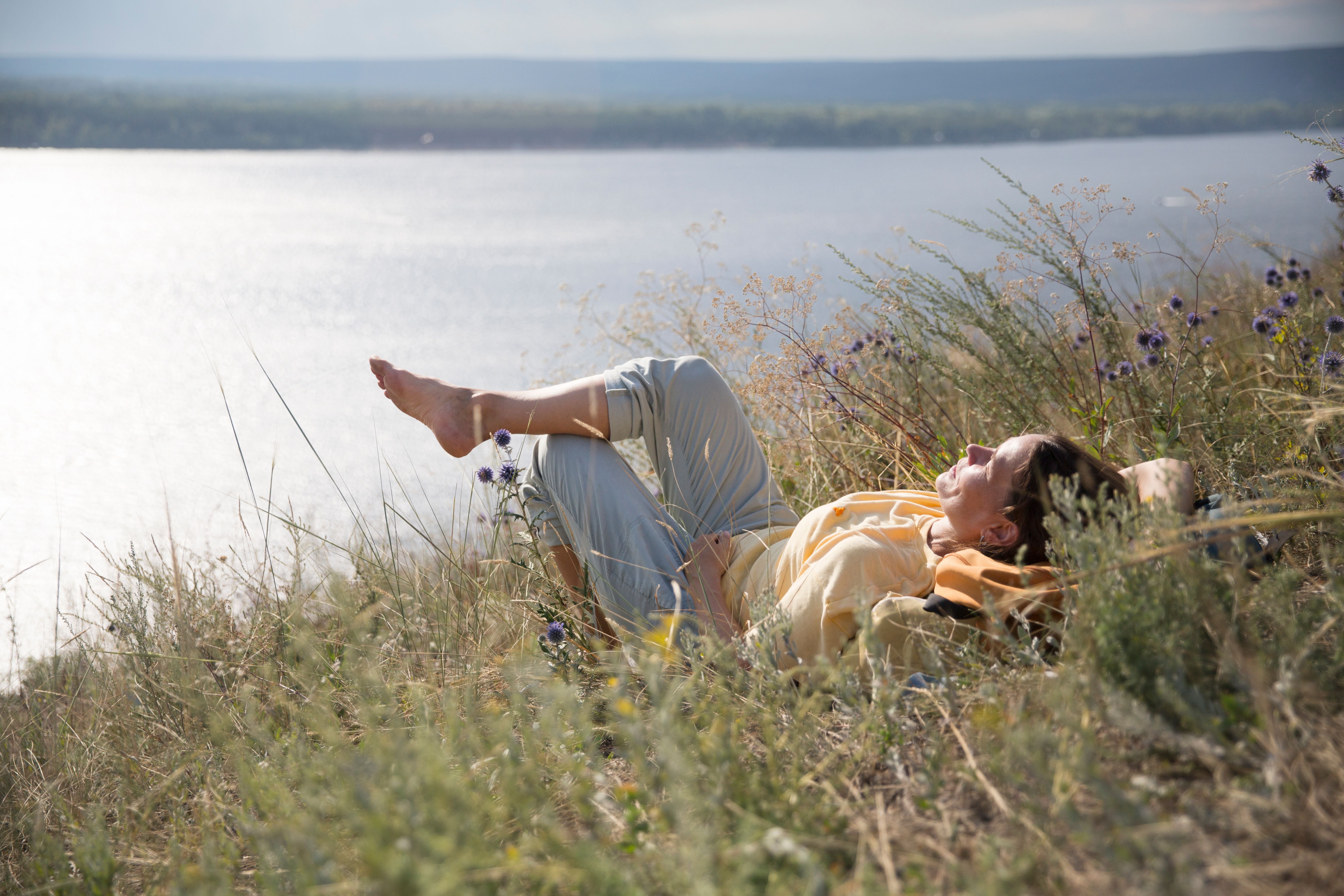 Frau liegt entspannt im Gras auf einem Hügel am See und genießt die Sonne.