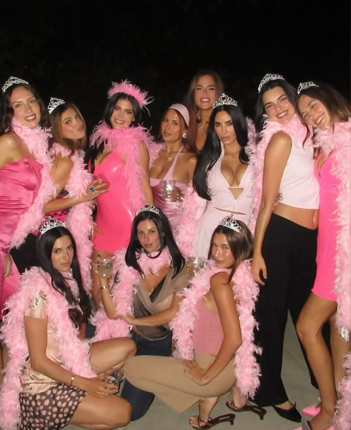 Mehrere Frauen in pinker Kleidung und mit Tiara posieren fröhlich für ein Gruppenfoto bei Nacht.