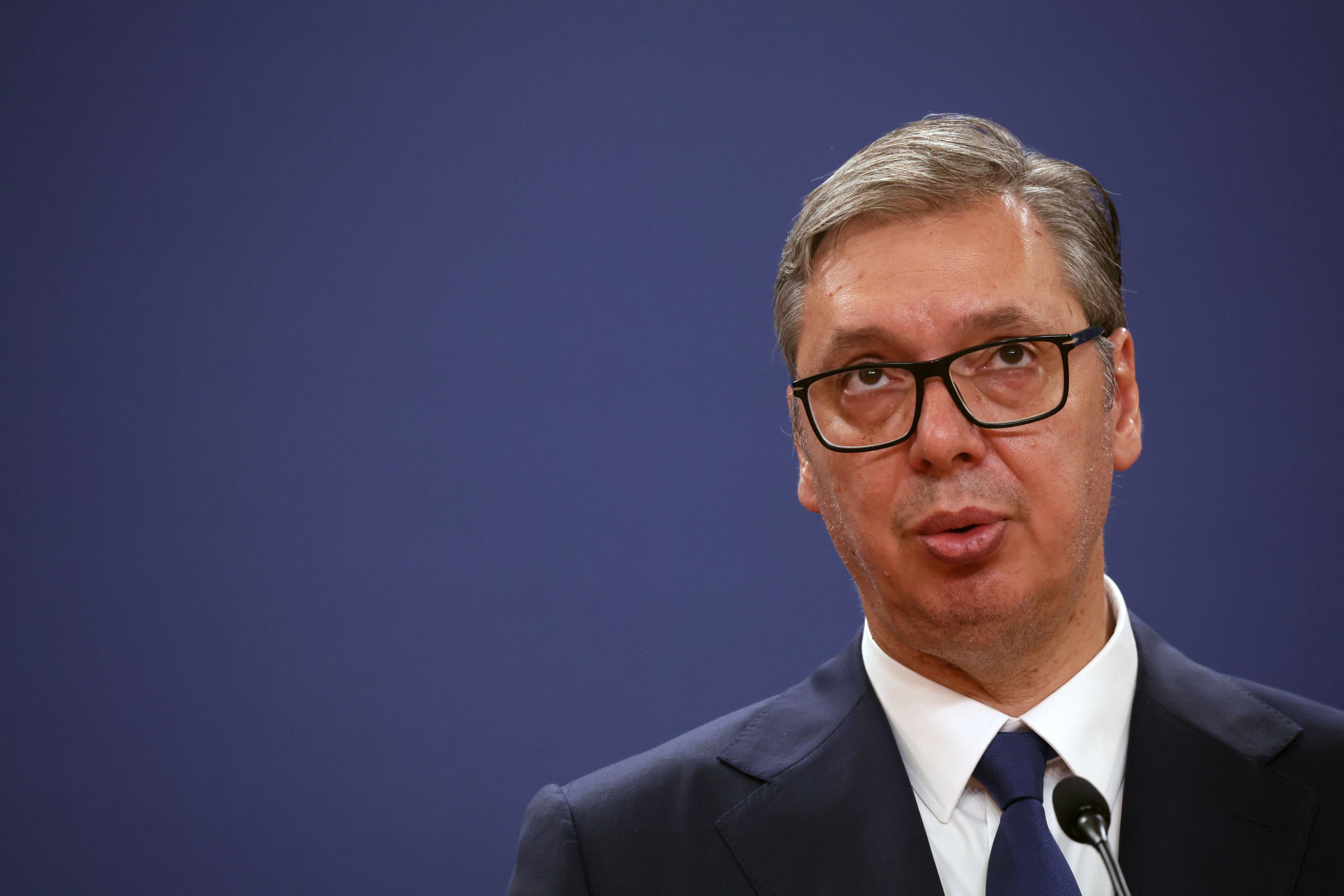 Ein Mann mit Anzug und Brille spricht vor Mikrofon auf einer Pressekonferenz, blauer Hintergrund.
