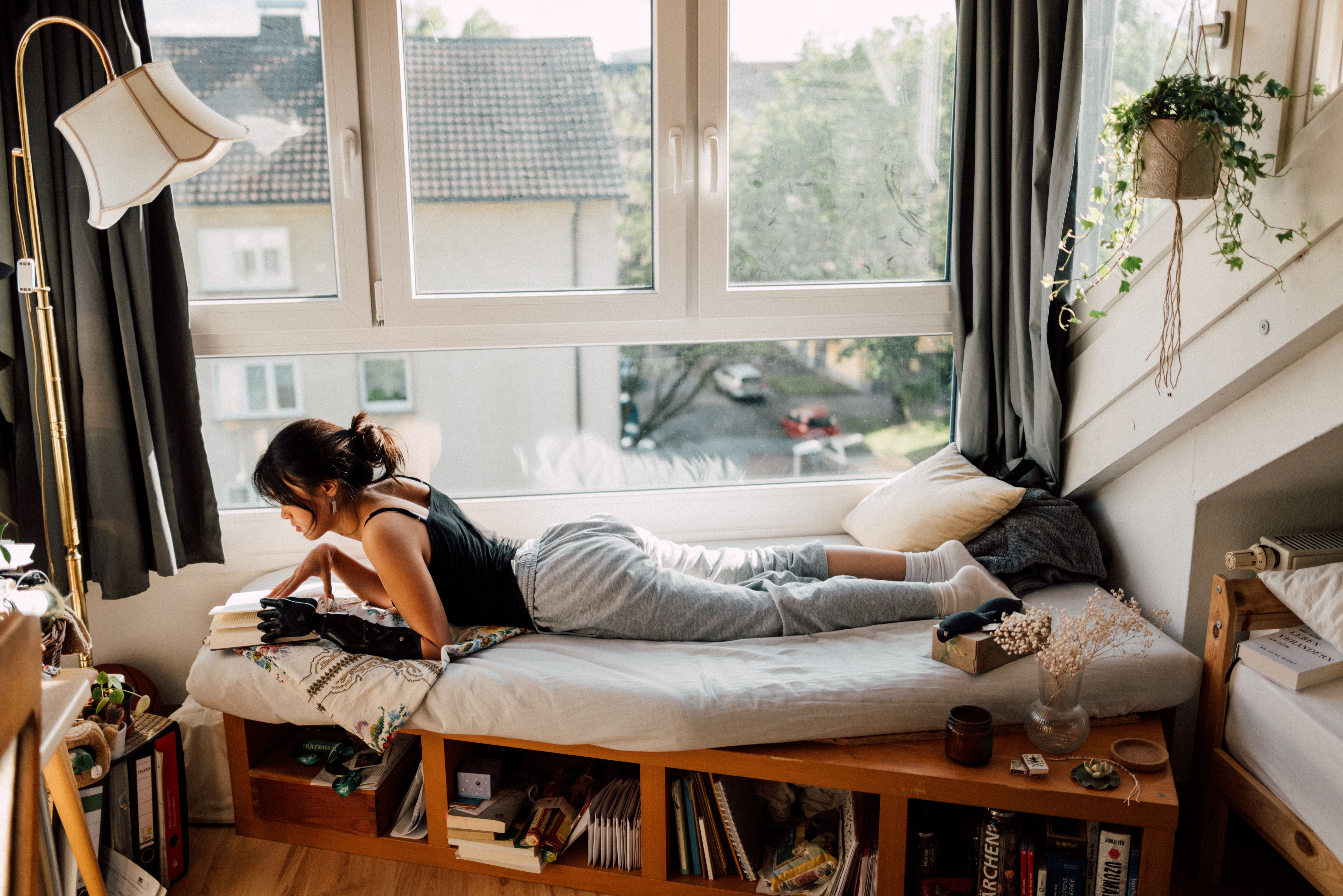 Frau liegt entspannt auf einem Bett und liest, Sonnenlicht fällt durch das Fenster ins gemütliche Zimmer.