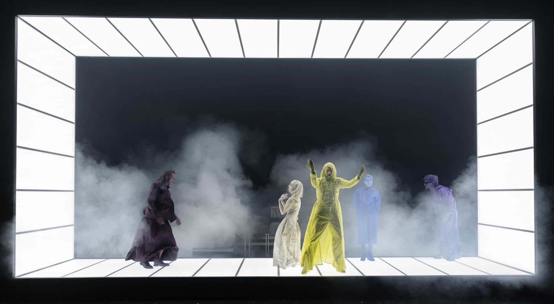Fünf Personen in farbigen Kostümen stehen auf einer beleuchteten Theaterbühne mit Nebel.