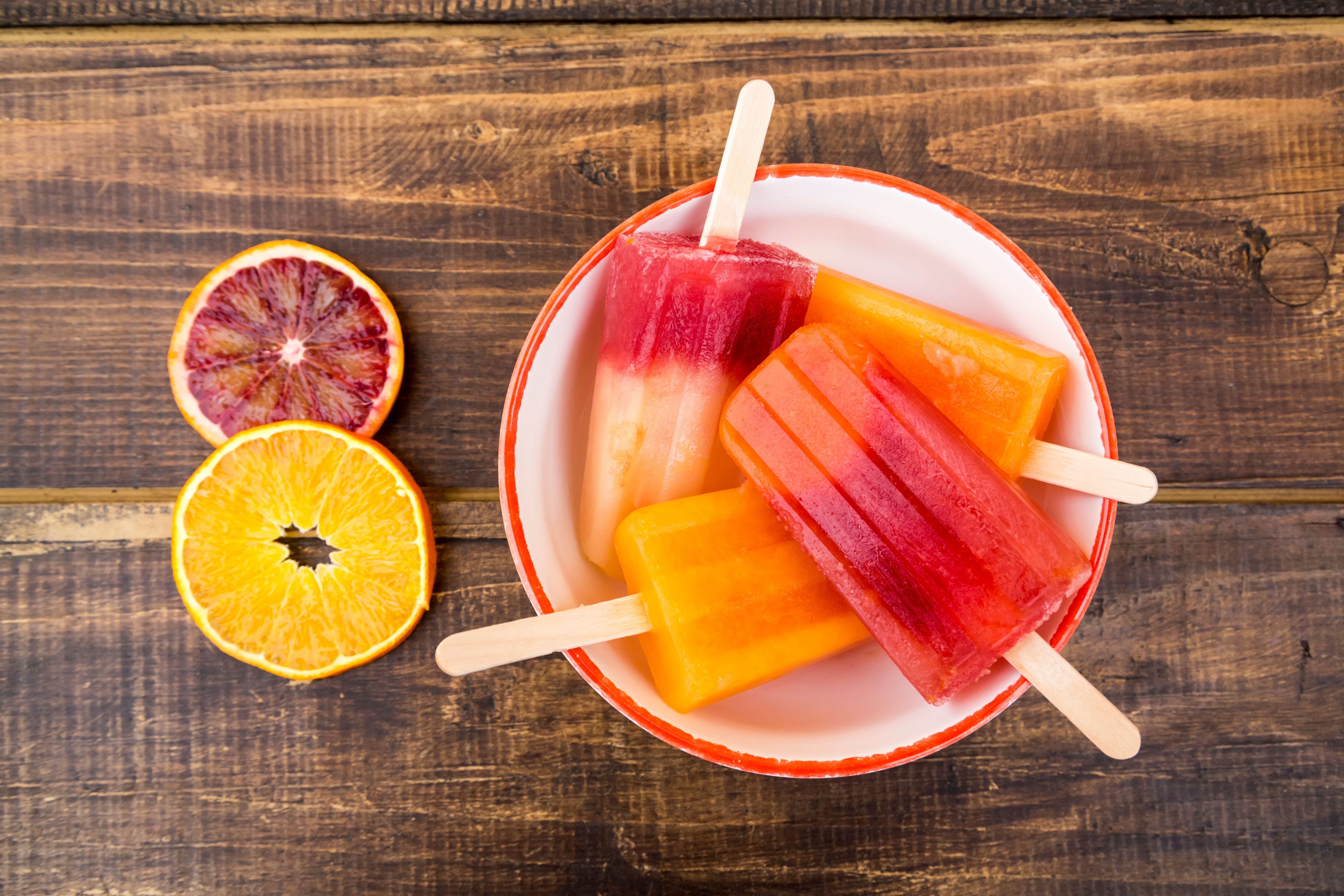 Drei Aperol-Spritz-Eis am Stiel in einer Schüssel, daneben zwei Orangenscheiben.
