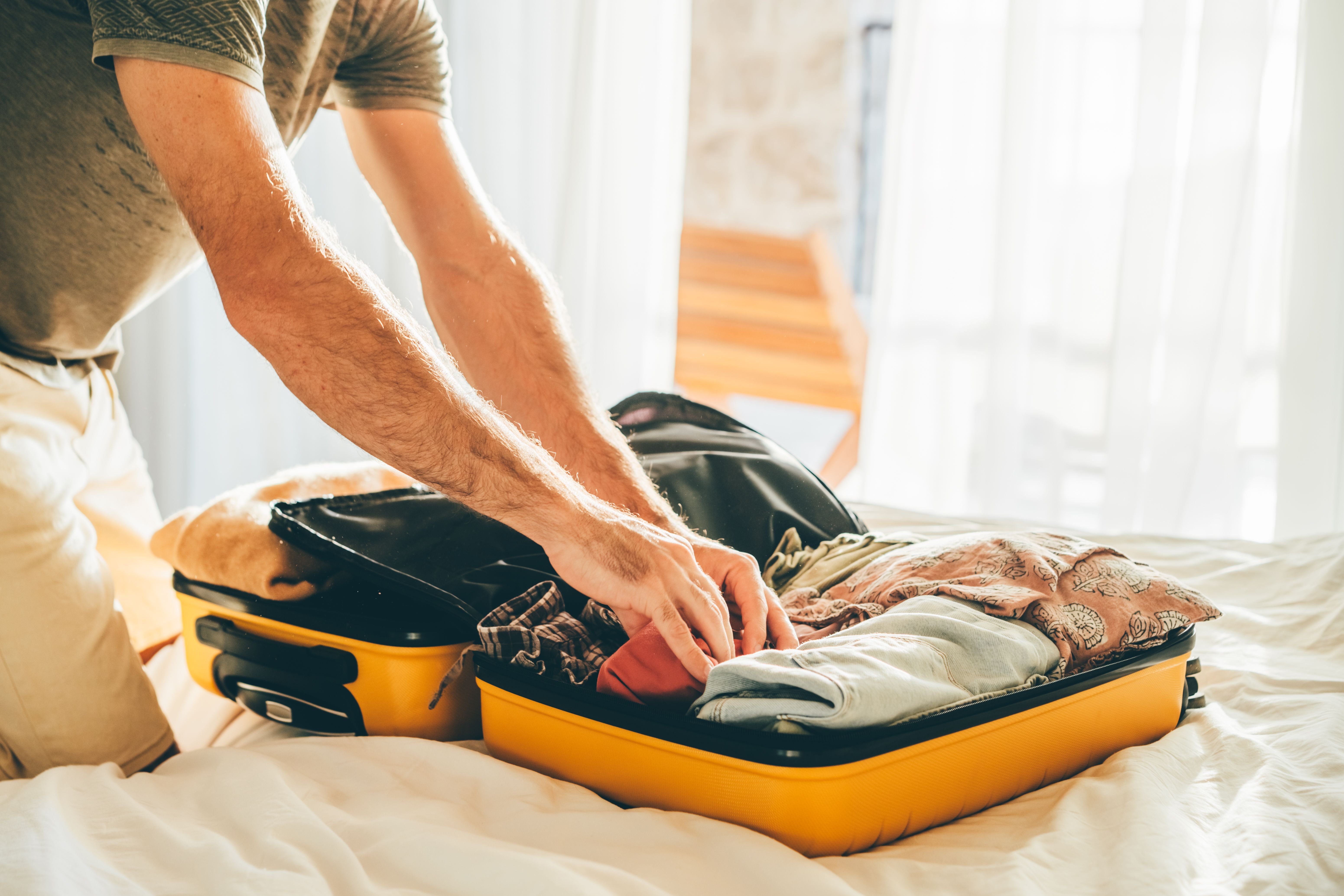 Mann packt Kleidung in einen gelben Koffer auf einem Bett in einem hellen Zimmer.