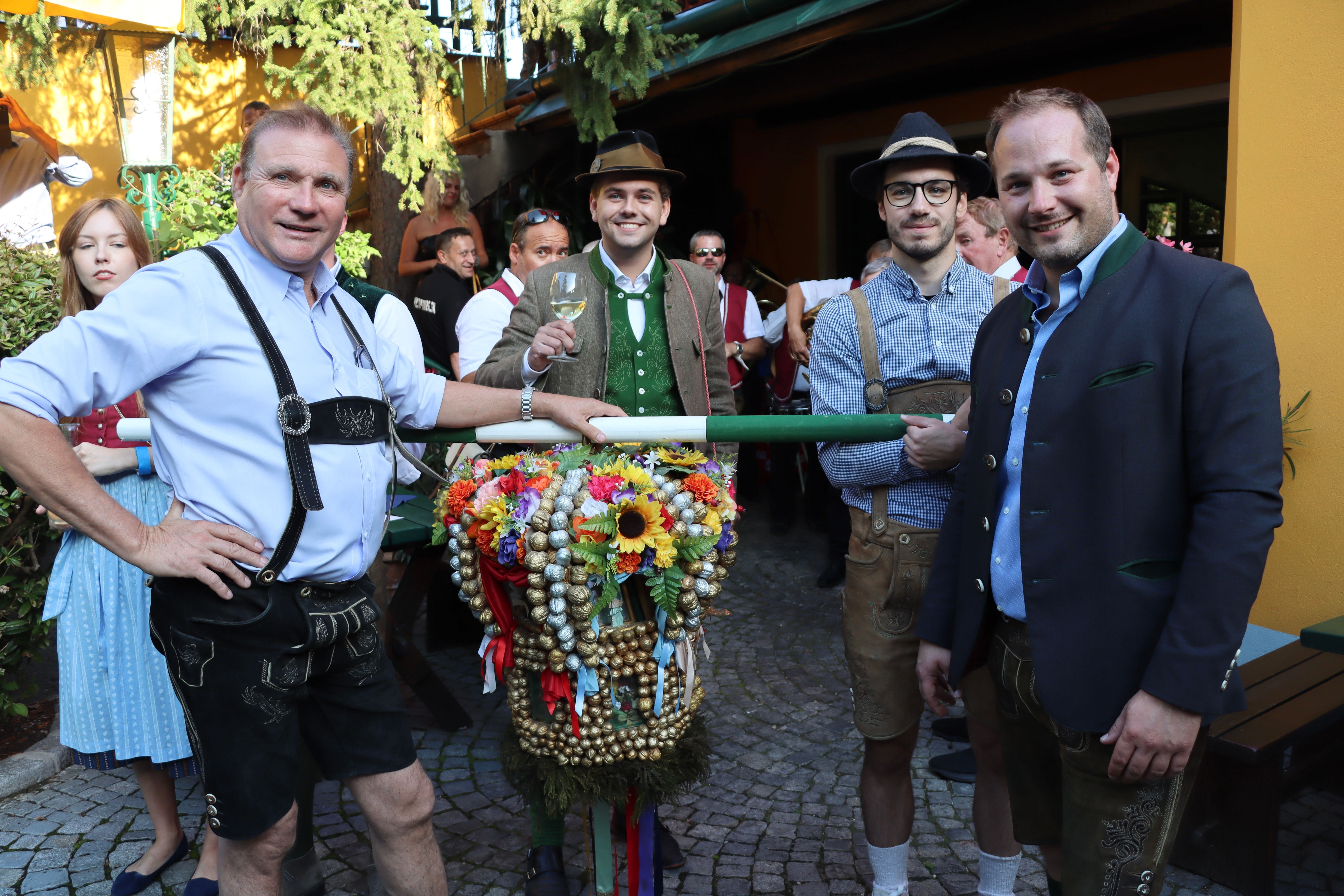 Vier Männer in traditioneller Tracht feiern draußen mit geschmücktem Kranz und Weinglas.