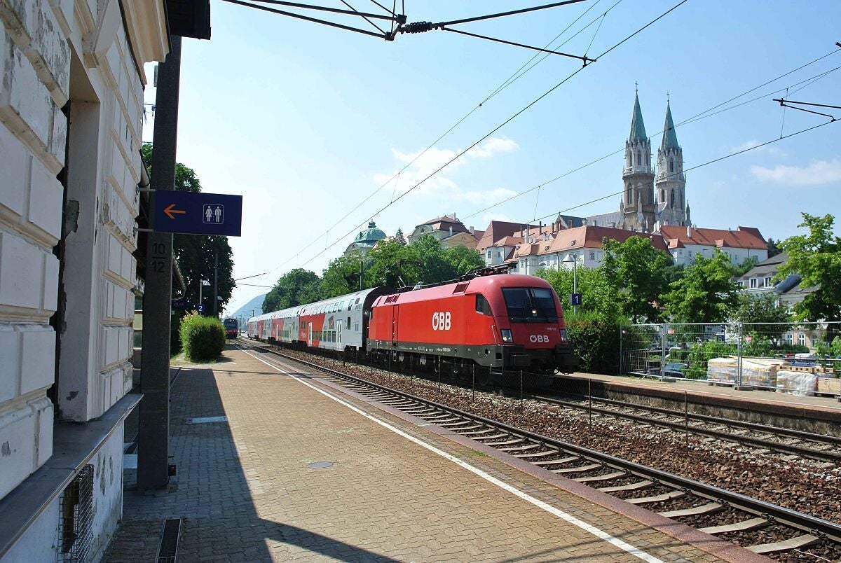 Ein roter ÖBB-Zug hält am Bahnhof, im Hintergrund das Stift Klosterneuburg.