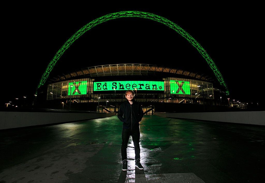 Ein Mann steht nachts vor dem grün beleuchteten Wembley-Stadion mit „Ed Sheeran X“-Schriftzug.