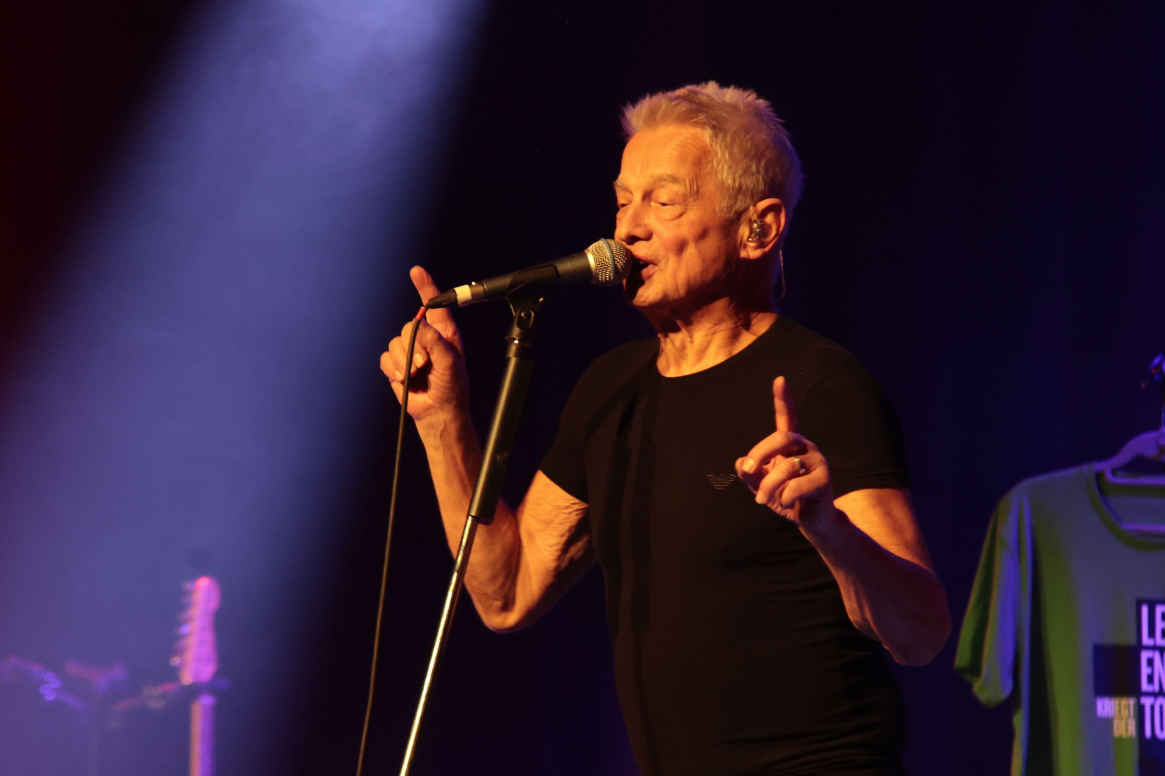 Ein älterer Mann singt auf einer Bühne in ein Mikrofon, im Hintergrund bunte Beleuchtung.