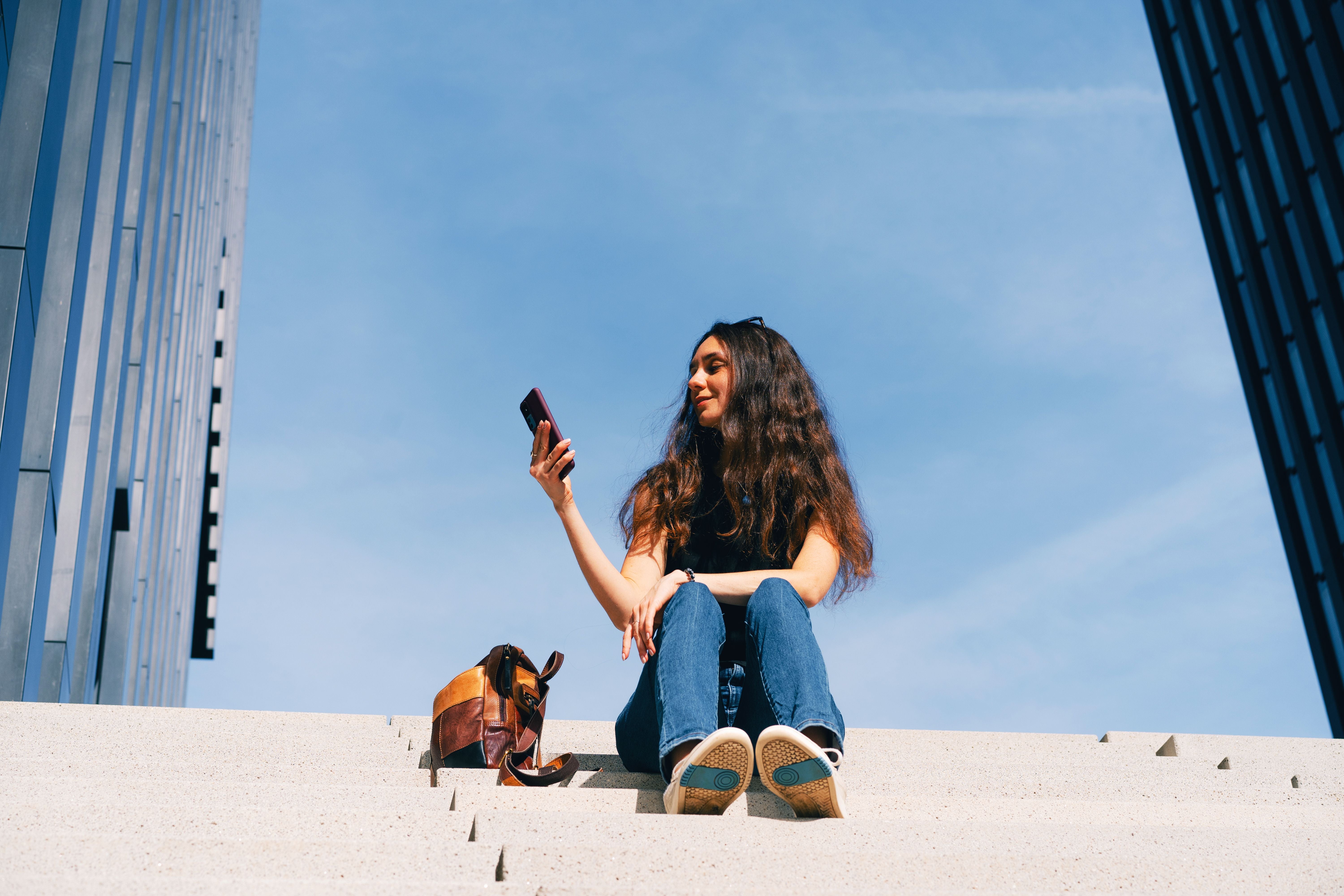 Junge Frau sitzt entspannt auf Treppen vor modernem Gebäude und schaut auf ihr Smartphone.