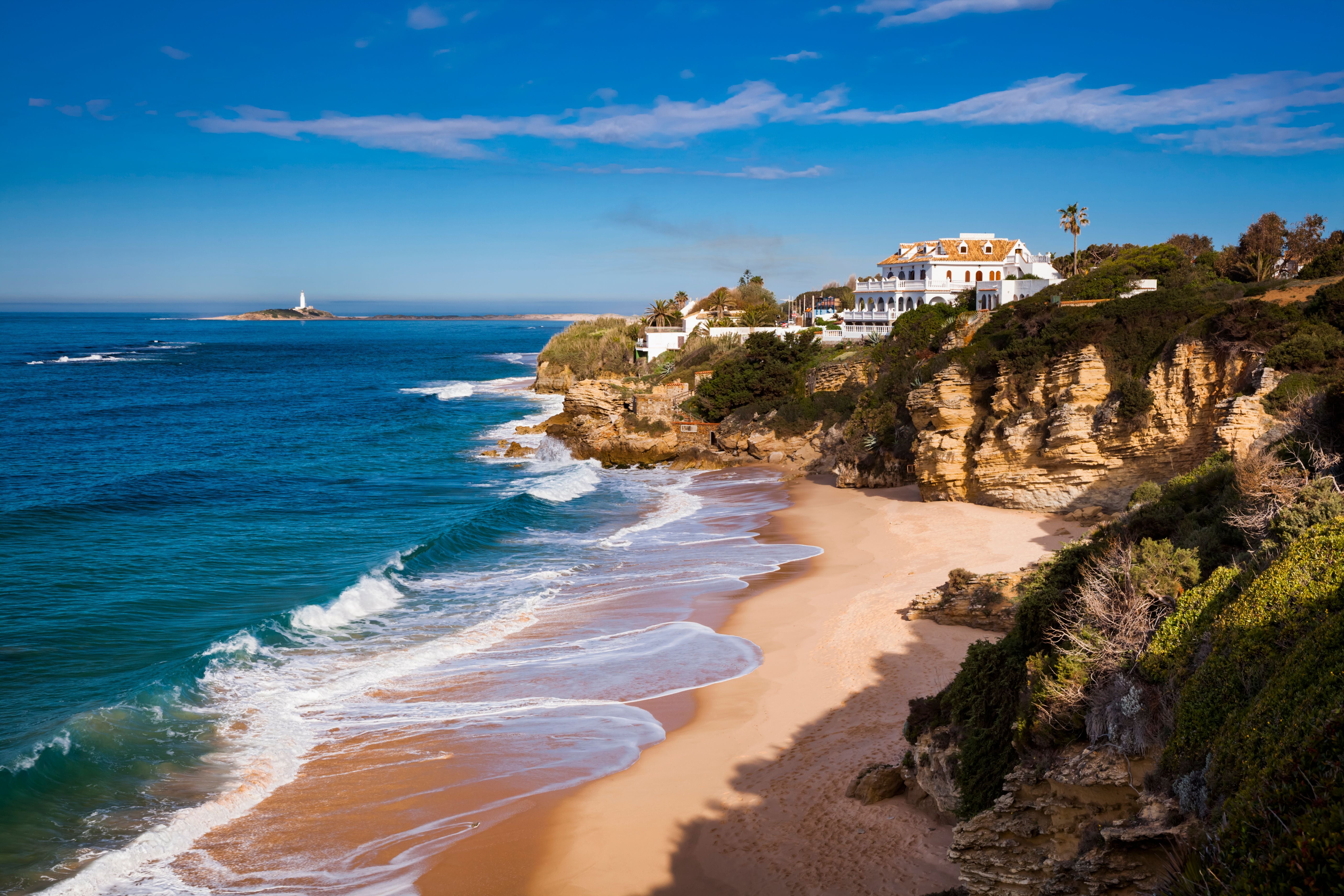 Sandstrand mit Felsen, weißem Gebäude und Leuchtturm im Hintergrund an der Costa de la Luz, Spanien.