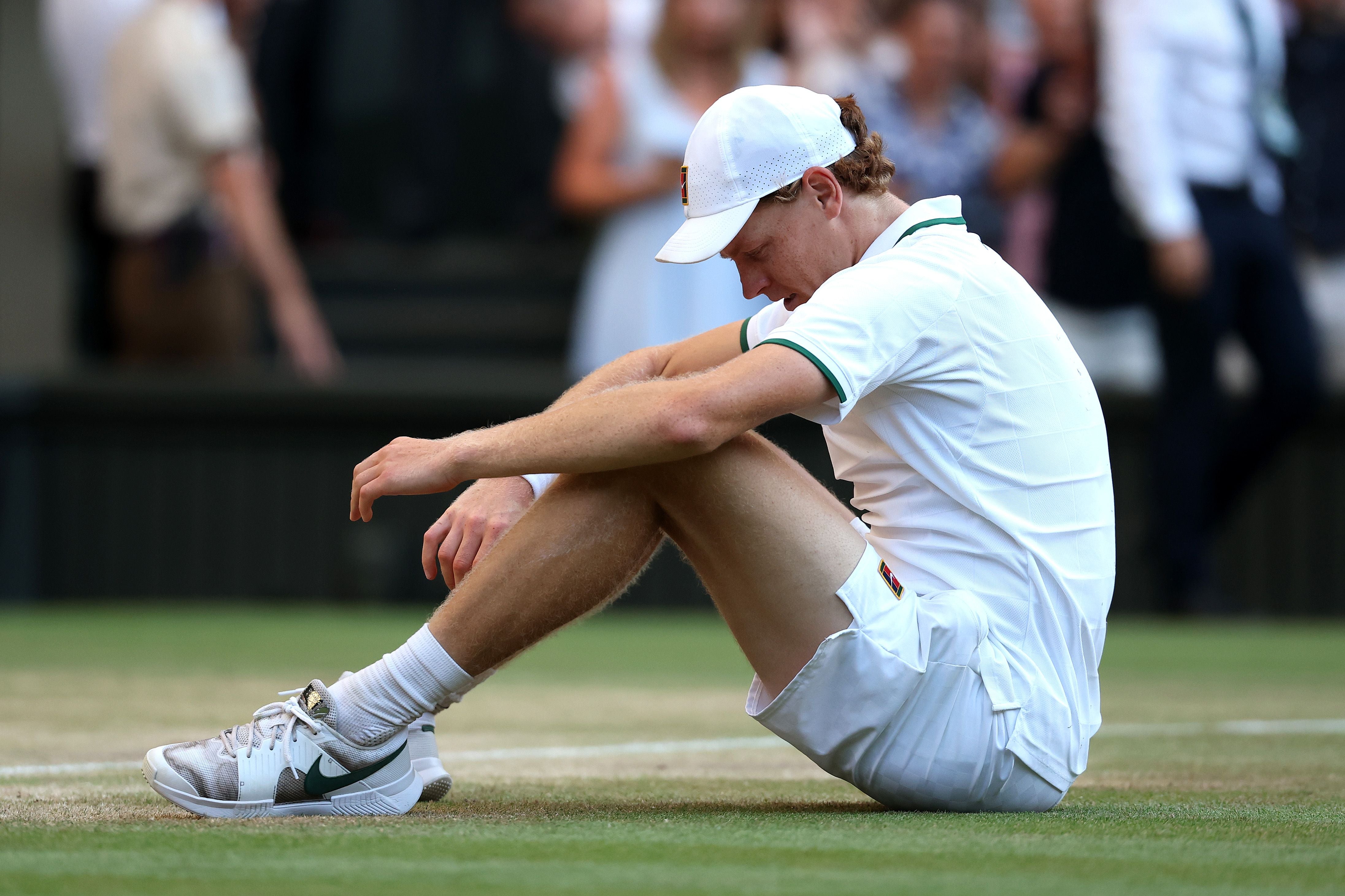 Ein Tennisspieler in weißem Outfit sitzt erschöpft auf dem Rasenplatz während eines Turniers.