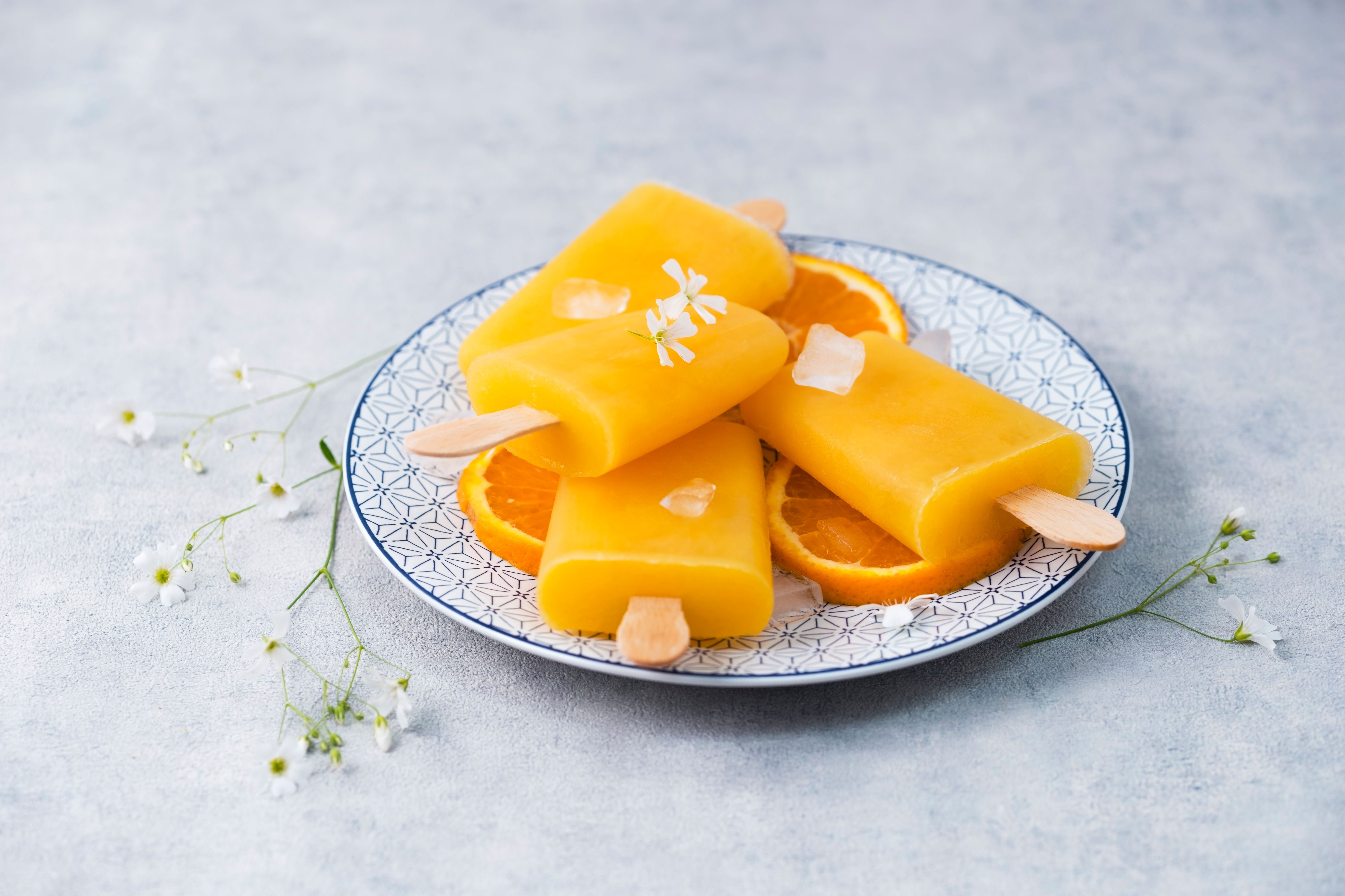 Drei Aperol-Spritz-Eis am Stiel auf Orangenscheiben und Eiswürfeln auf einem Teller.