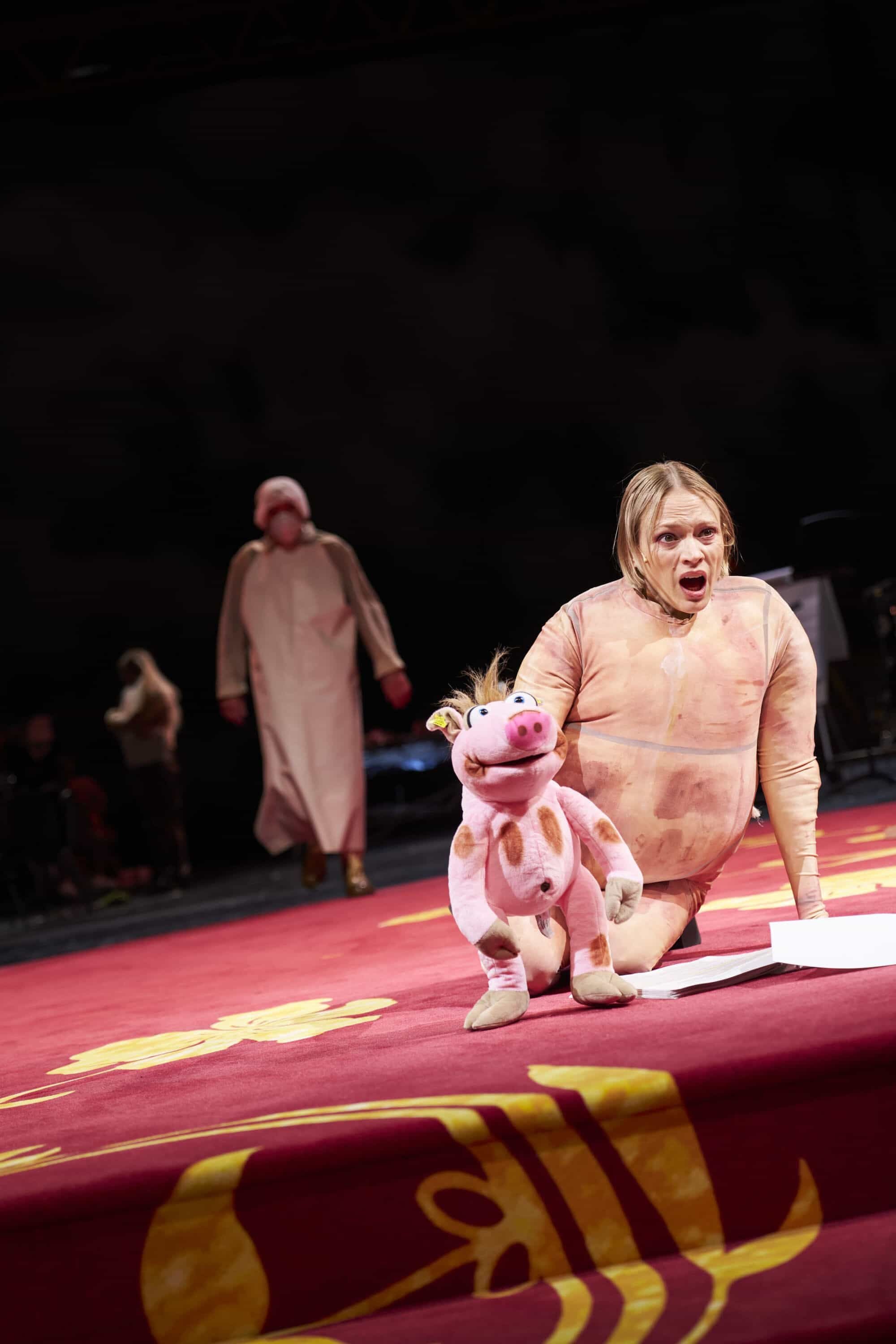 Schauspielerin im Tierkostüm kniet auf Bühne mit rosa Stoffschwein bei den Salzburger Festspielen.