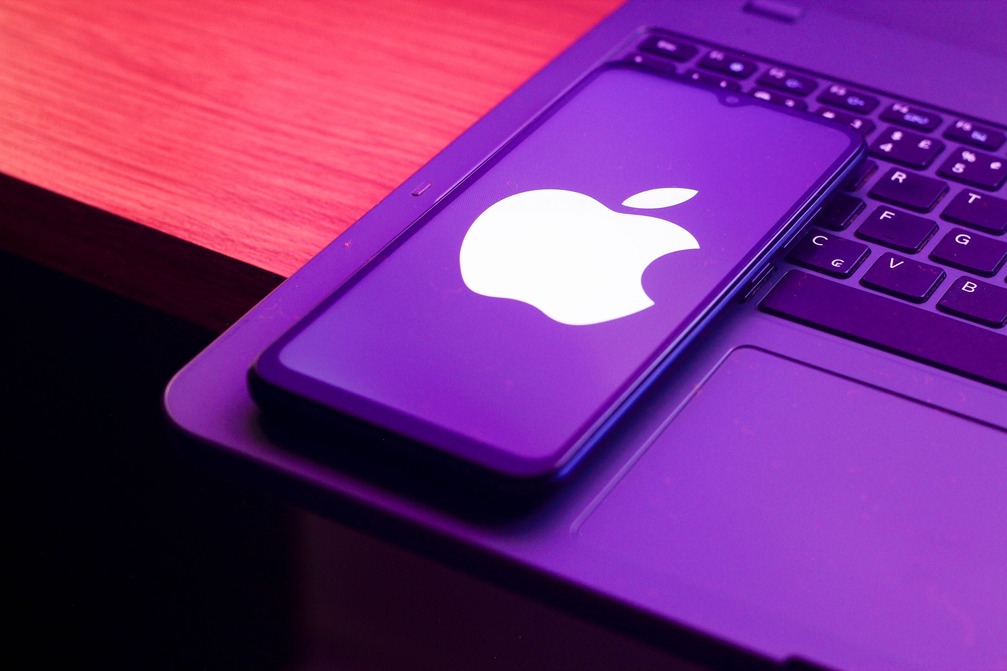 Ein Smartphone mit Apple-Logo liegt auf einer Laptop-Tastatur in violettem Licht.
