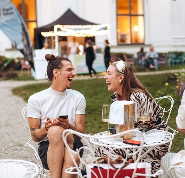 Zwei junge Menschen lachen gemeinsam an einem Tisch im Garten des Palais Auersperg.