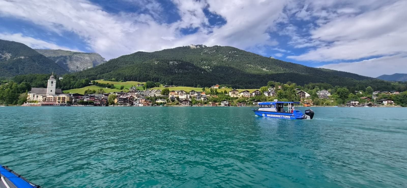 Blauer Rettungsboot auf dem Wolfgangsee vor einem Dorf mit Kirche und grünen Bergen im Hintergrund.