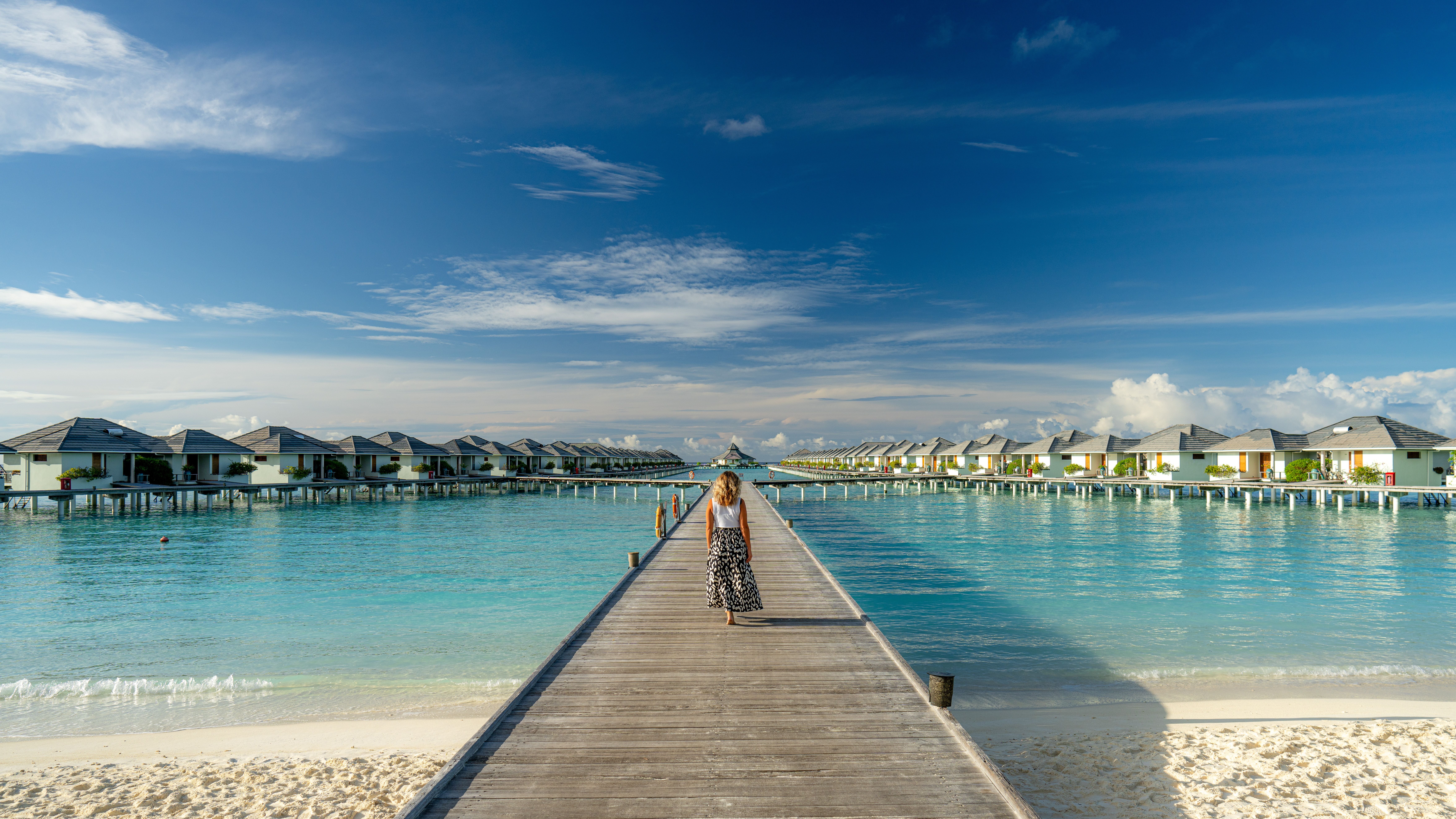 Frau geht auf Steg über türkisfarbenes Wasser, umgeben von Bungalows, unter blauem Himmel.