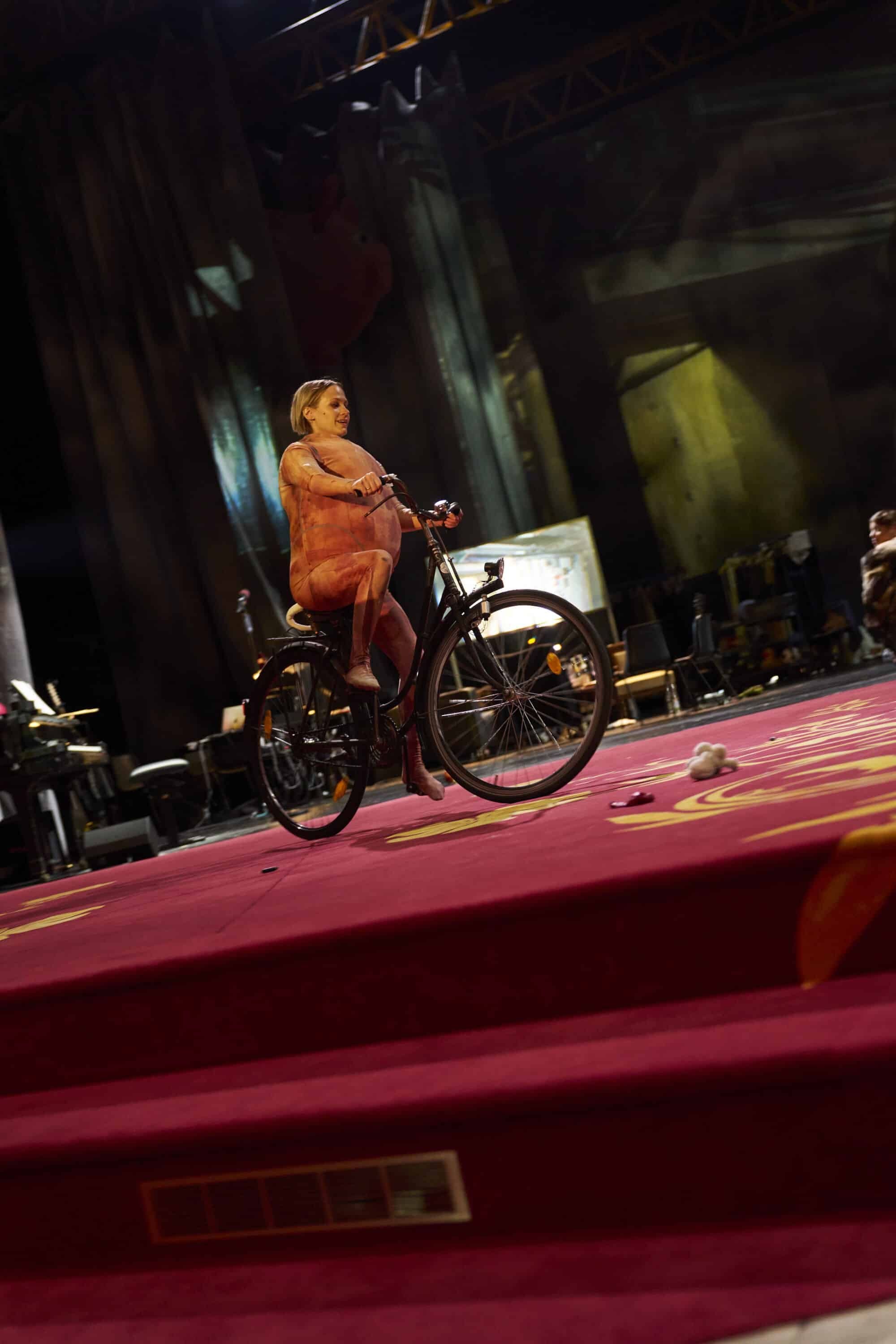 Eine Frau im hautfarbenen Kostüm fährt auf einem Fahrrad über eine rote Bühne im Theater.
