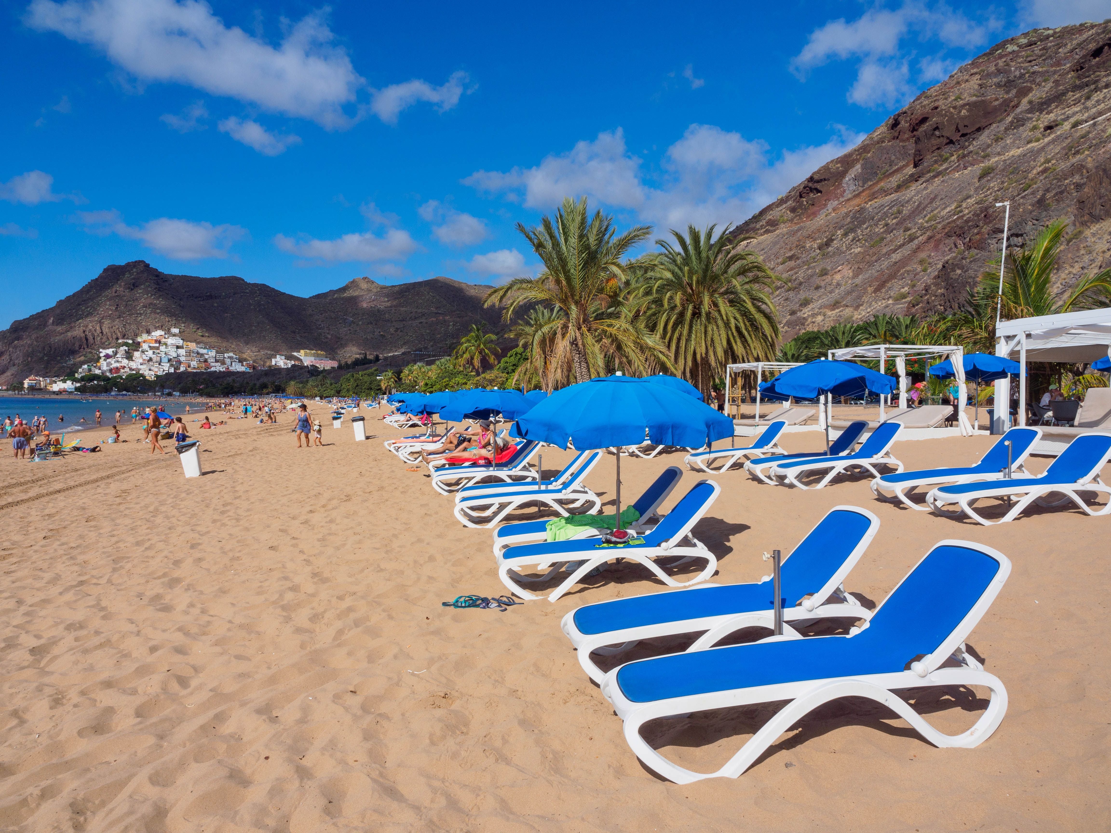 Liegestühle und blaue Sonnenschirme am Sandstrand mit Palmen und Bergen im Hintergrund.
