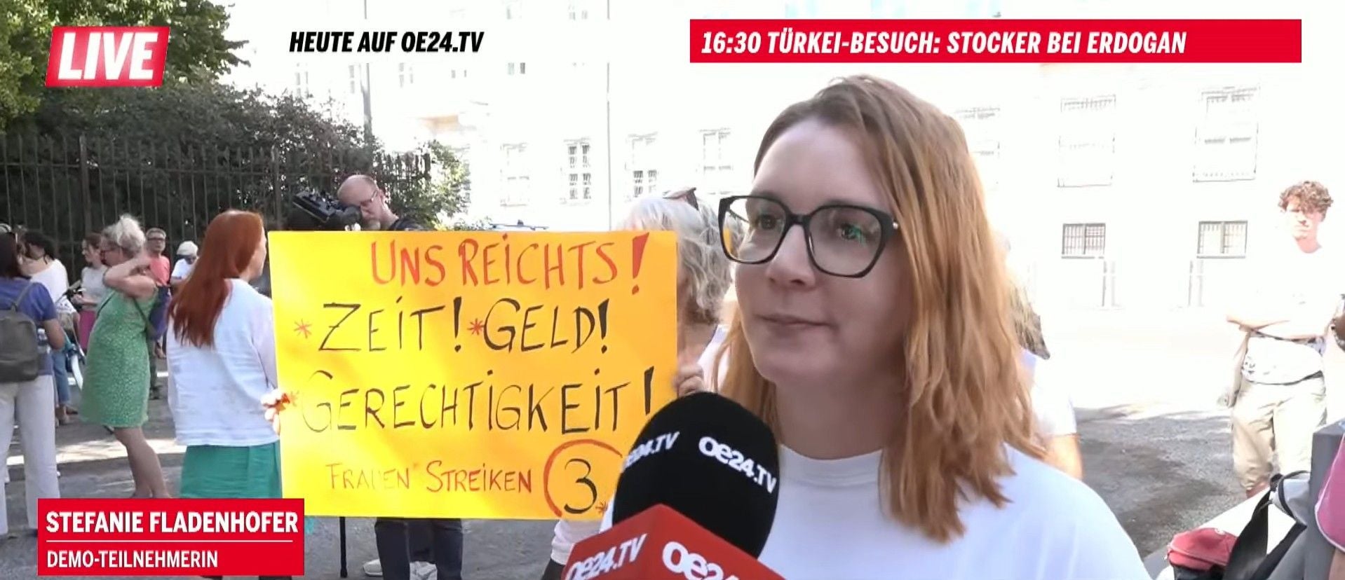 Eine Frau wird interviewt, im Hintergrund hält jemand ein gelbes Protestplakat hoch.
